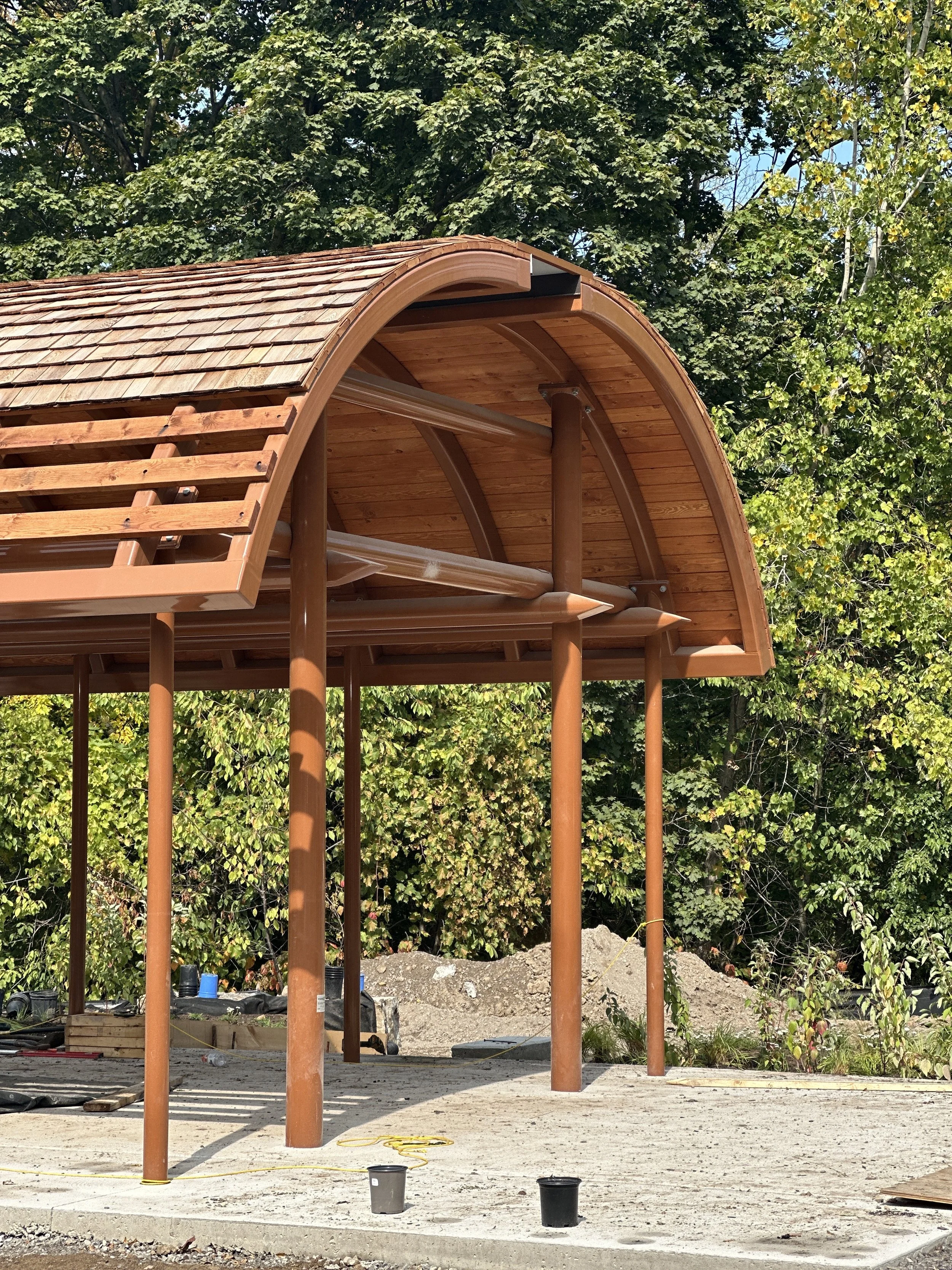 Under construction wooden structure with a curved roof, supported by brown poles, on a concrete foundation, surrounded by trees and construction materials.