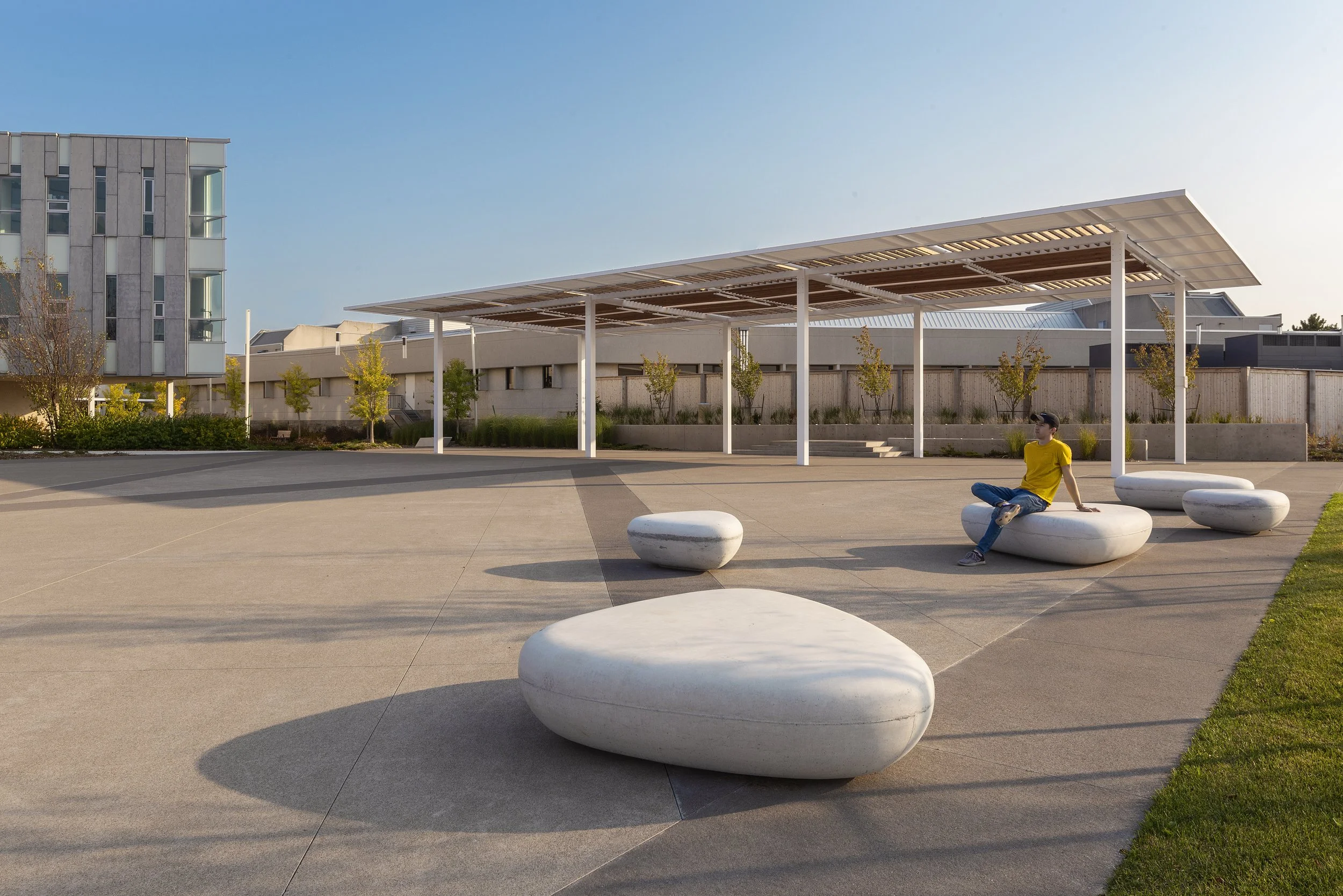 Open plaza with large white stones used as seating, a young man in a yellow shirt sitting on one of the stones, small trees along the background, a modern building to the left, and a shaded structure overhead, all under a clear blue sky.
