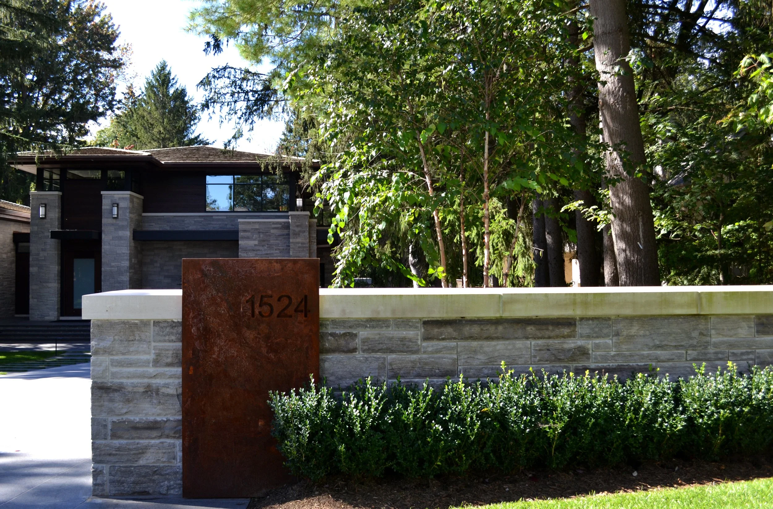 Modern house with a stone and concrete fence, house number 1524, surrounded by green bushes and tall trees, sunny day.