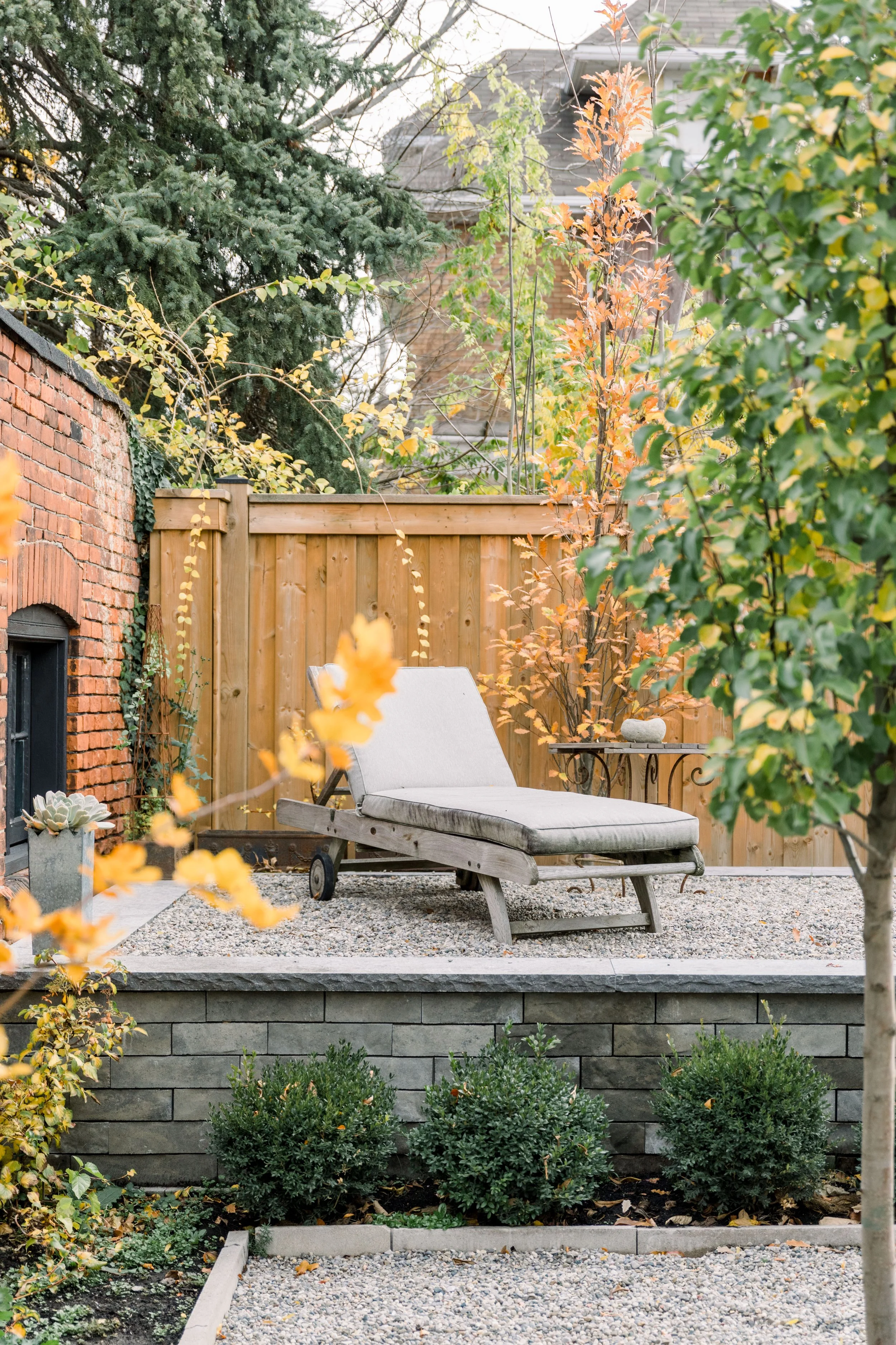 A cozy backyard patio with a lounge chair, trees with orange and green leaves, a brick wall, and a wooden fence, all set on a gravel and stone surface.
