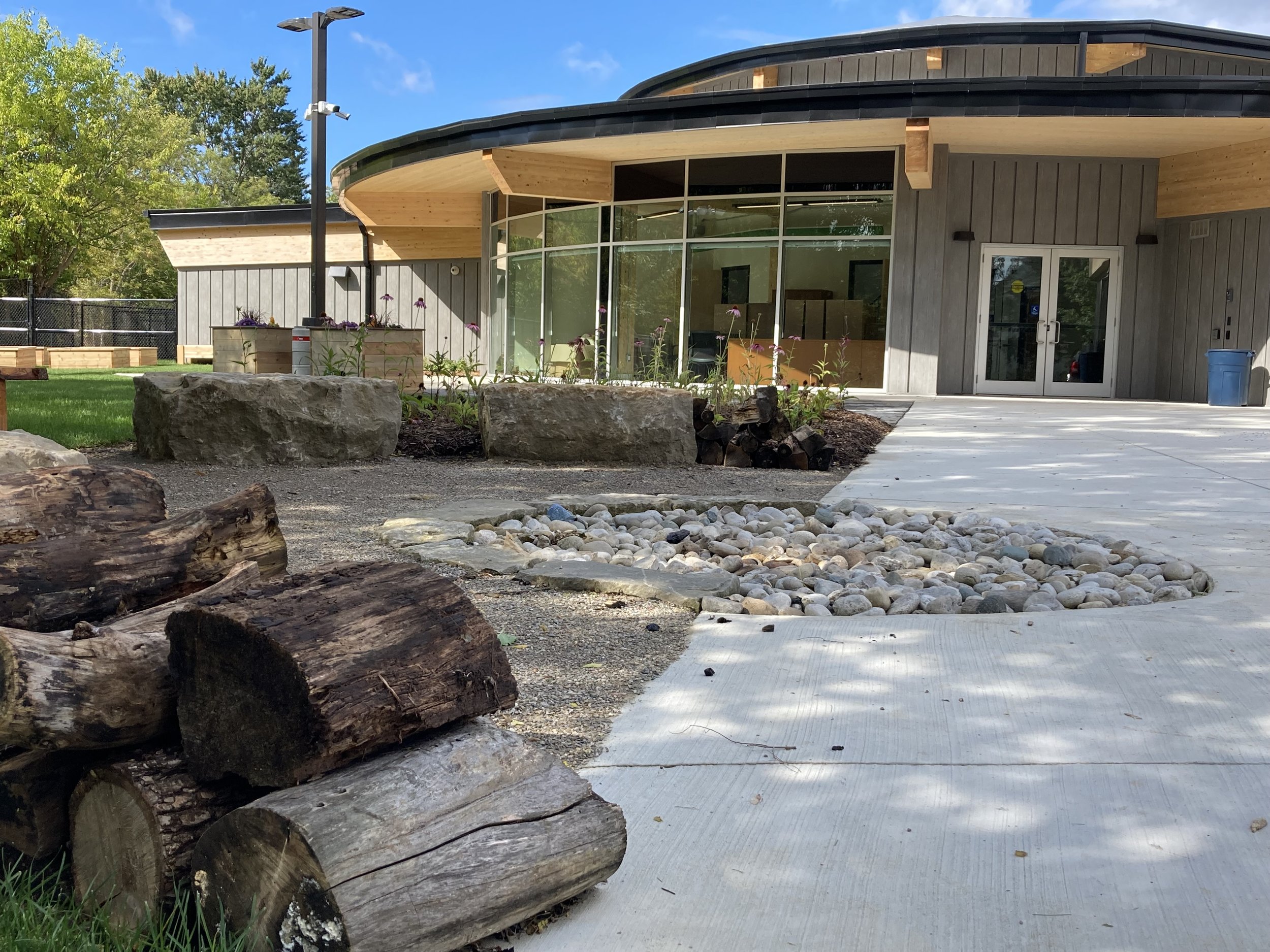 Modern building with large glass windows, wooden accents, and a concrete walkway, surrounded by a green lawn and landscaping with rocks and logs.