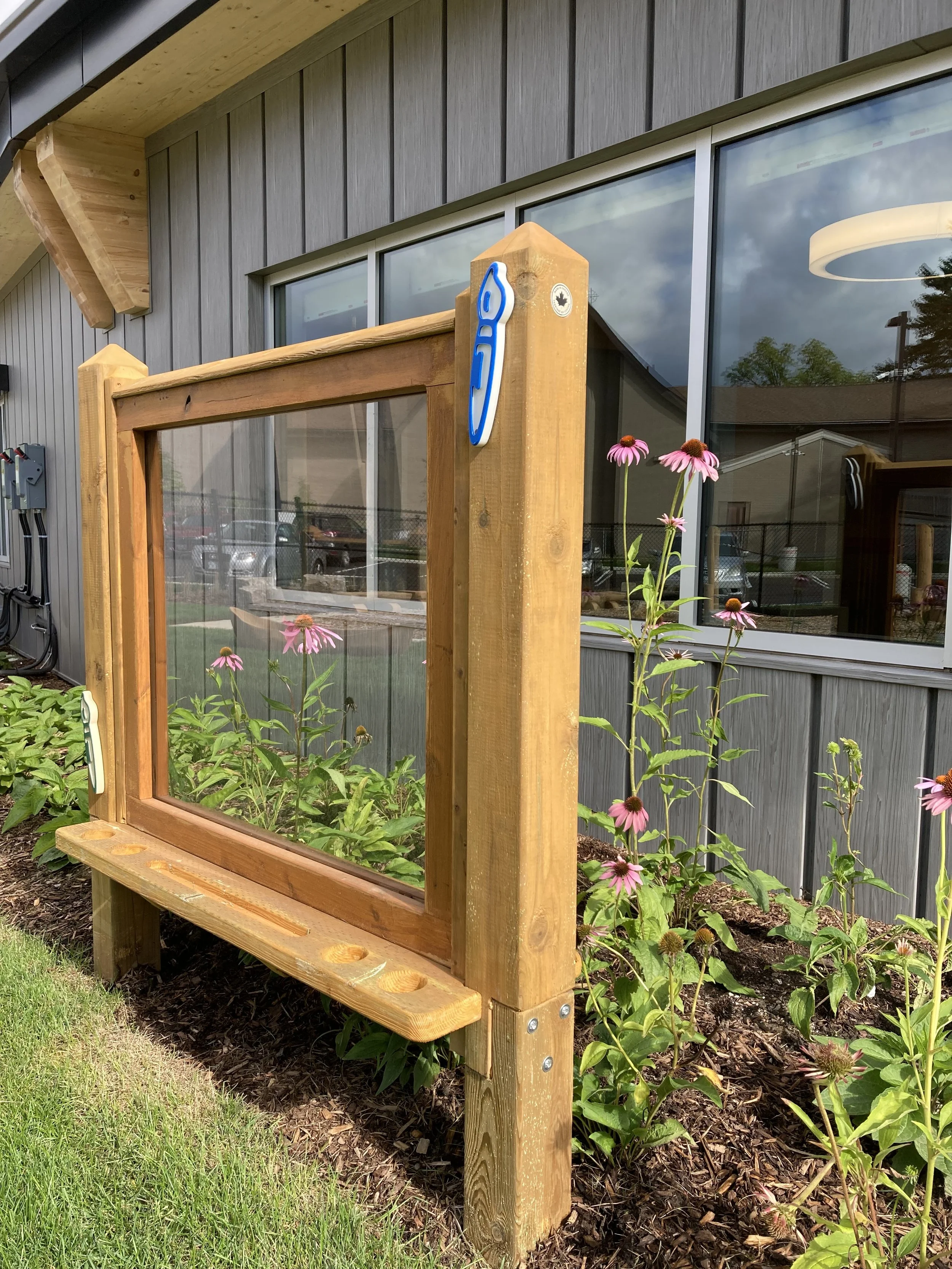 A wooden outdoor display case with a glass front, mounted on a garden bed with pink coneflowers, in front of a gray building with large windows. There are a couple of stickers on the corner of the display case, one of a blue utility knife and one of 