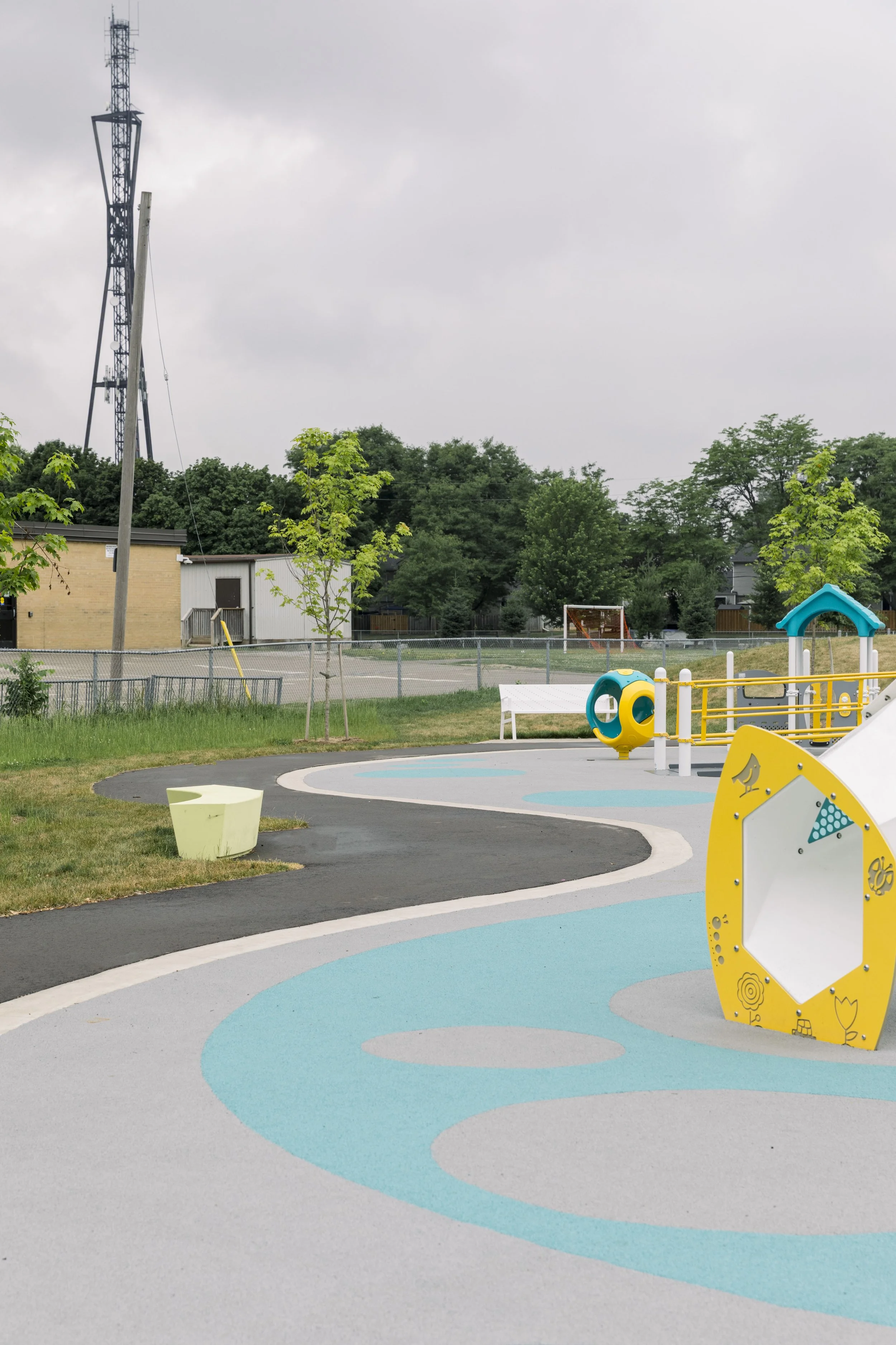 Empty playground with colorful rubberized flooring, small play structures, and surrounding trees under cloudy sky.
