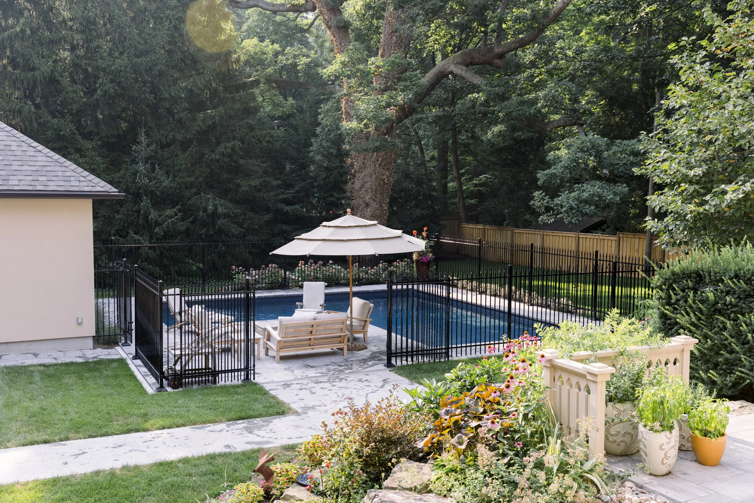 A backyard with a swimming pool surrounded by a black metal fence, potted plants, and vibrant flowers. There are outdoor chairs and a large white patio umbrella, with trees and greenery in the background.