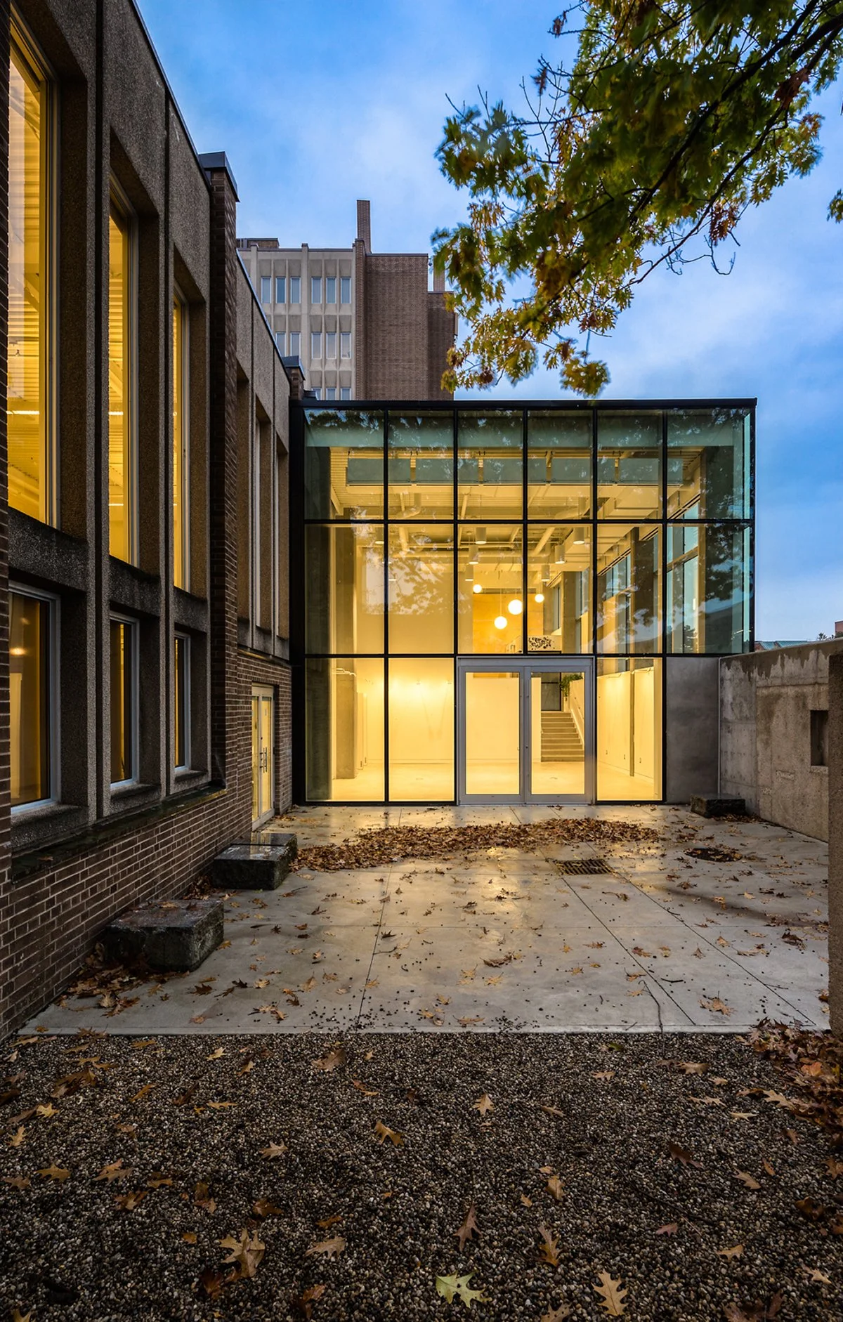 A modern building with large glass windows and a door, illuminated from within, situated in an urban area with some trees and multi-story buildings in the background in the evening.