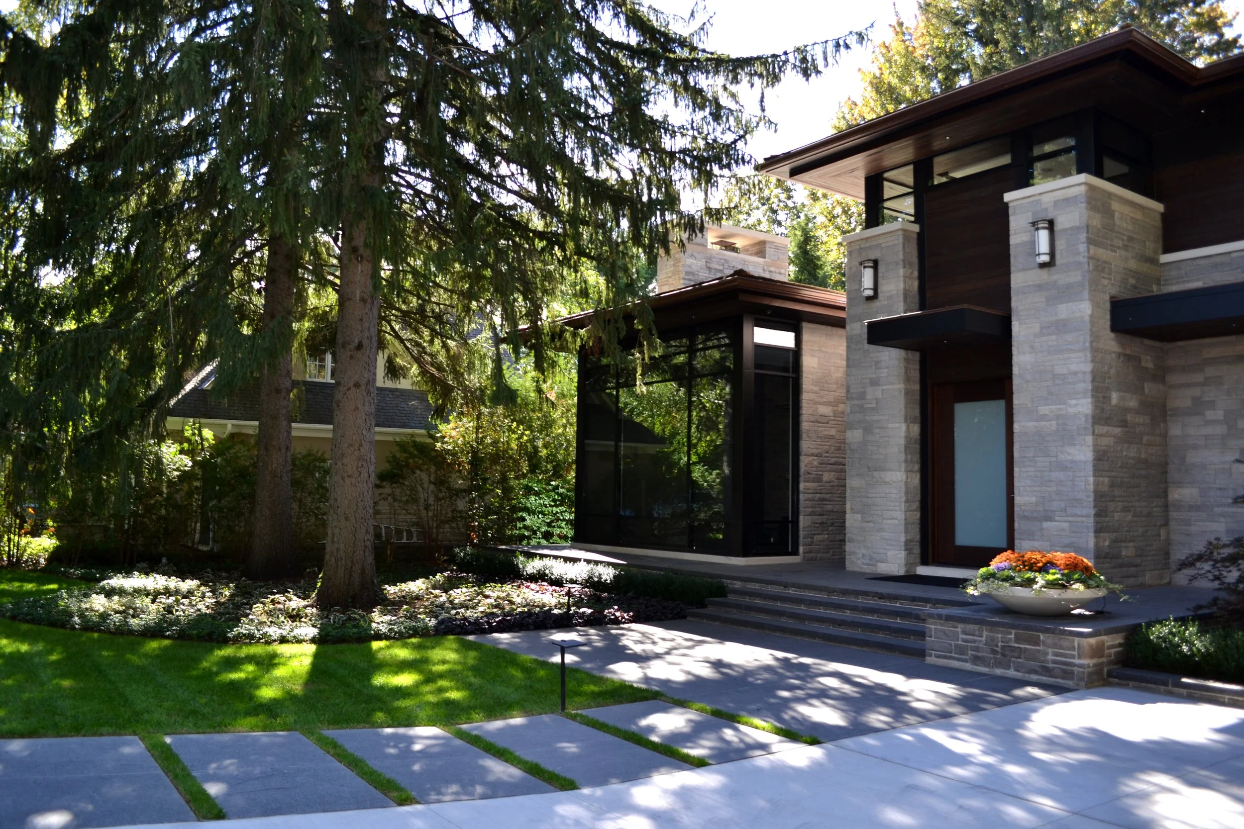 Modern house with stone exterior, glass entryway, and flower pot in front, surrounded by trees and greenery.
