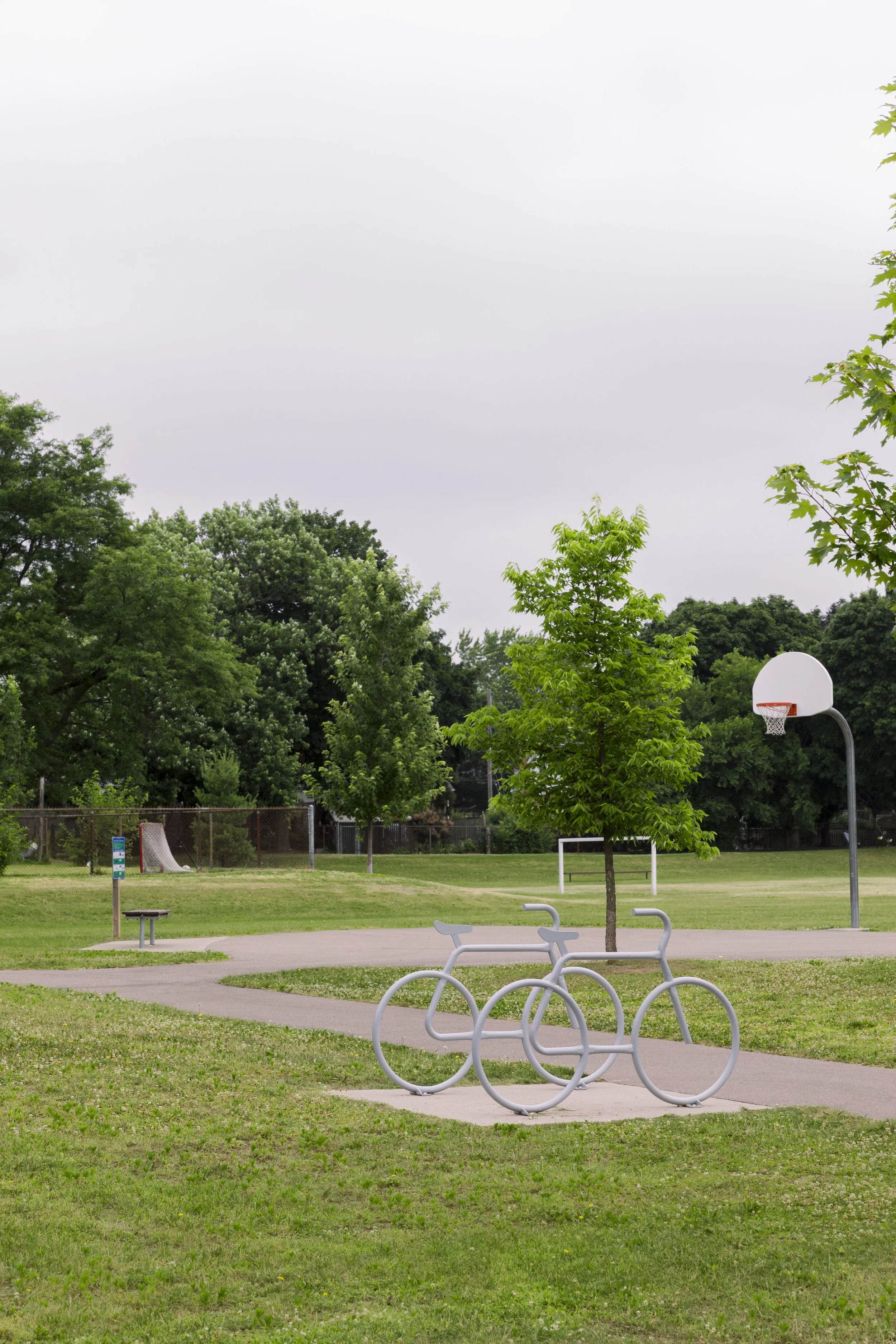 A park scene with green grass, trees, a basketball hoop, and bike racks shaped like bicycles.