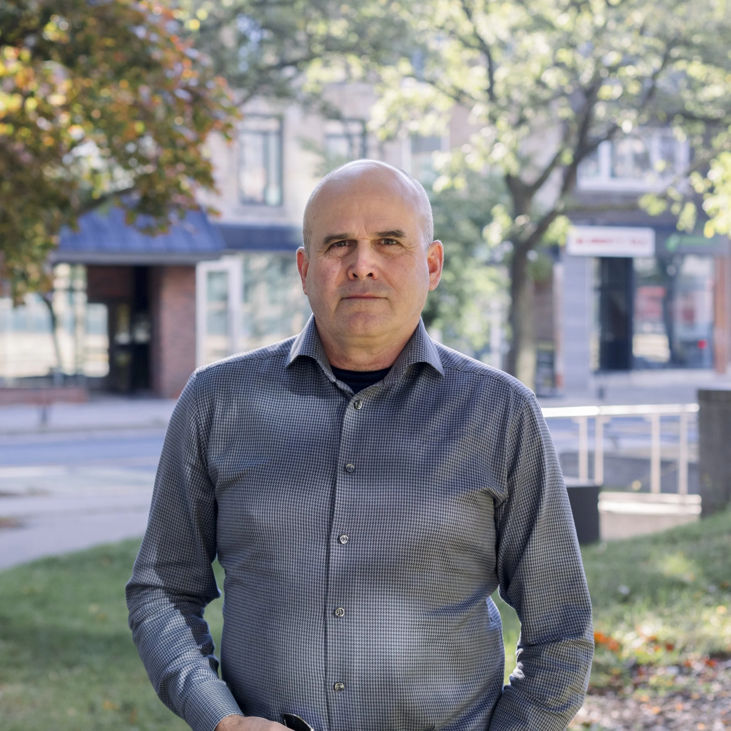 A middle-aged man with a serious expression standing outdoors in a city park during daytime. He is wearing a gray, patterned button-up shirt and has a bald head. Behind him are trees with green leaves, a sidewalk, and buildings with storefronts.