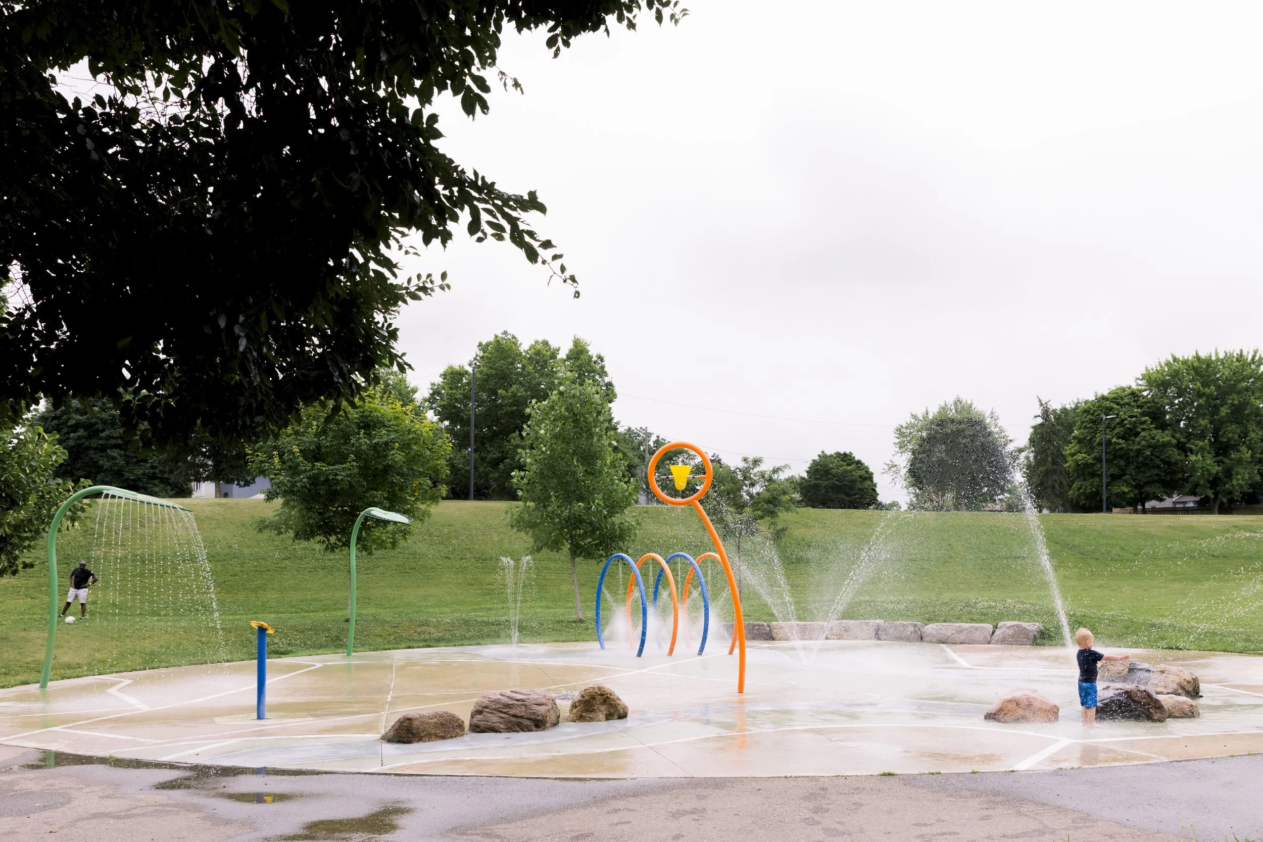 A children’s splash pad with water fountains, rocks, and play structures in a park with green trees and grass, and a cloudy sky in the background.