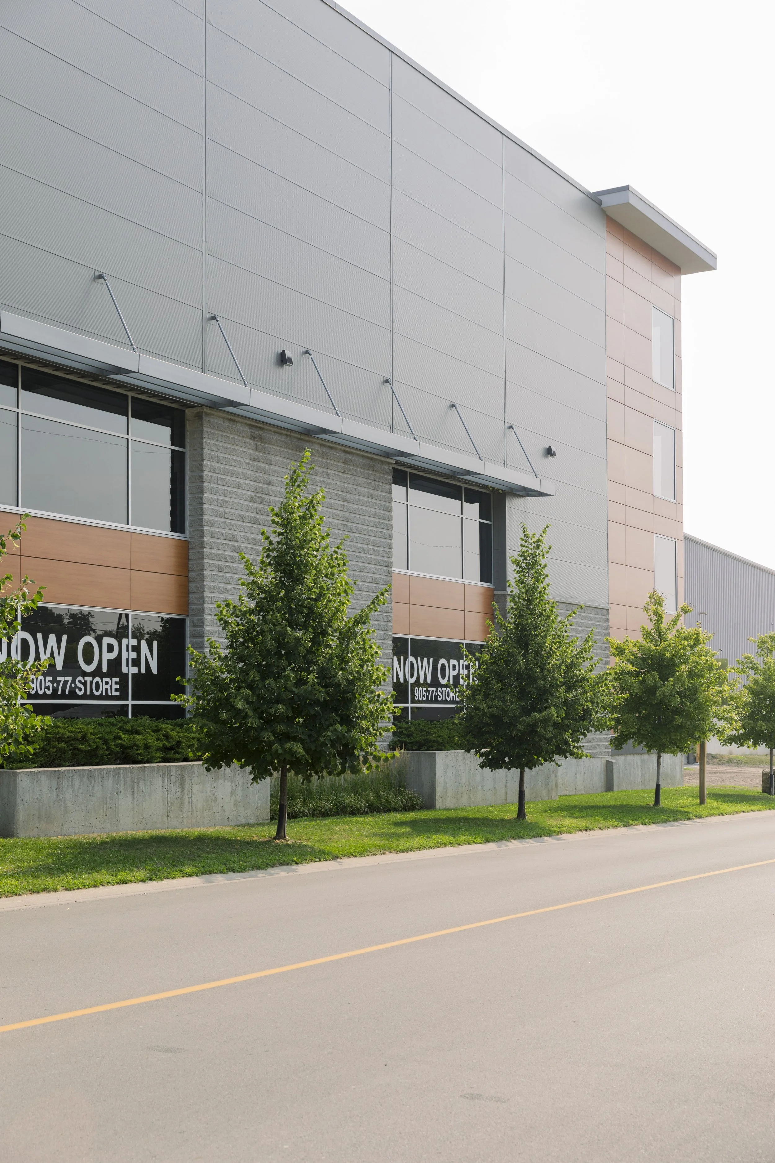 Exterior view of a modern commercial building with large windows and 'Now Open' signs, surrounded by green trees and a paved street.