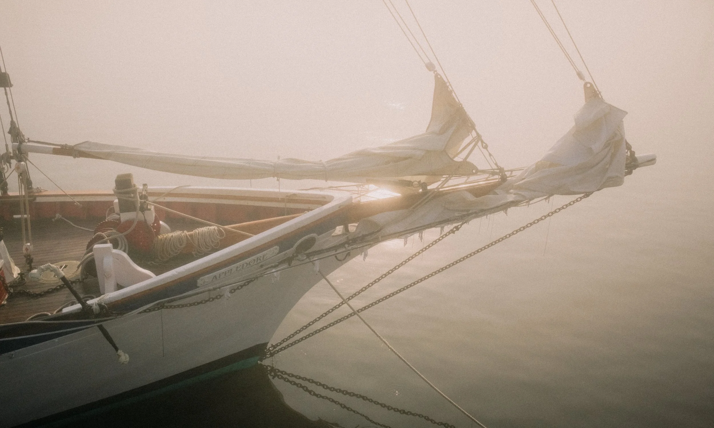 Camden, Maine. A sailboat docked on calm water during sunrise or sunset, with its sails furled and chains securing it in place, surrounded by mist or fog. Virginia Travel Photographer. 
