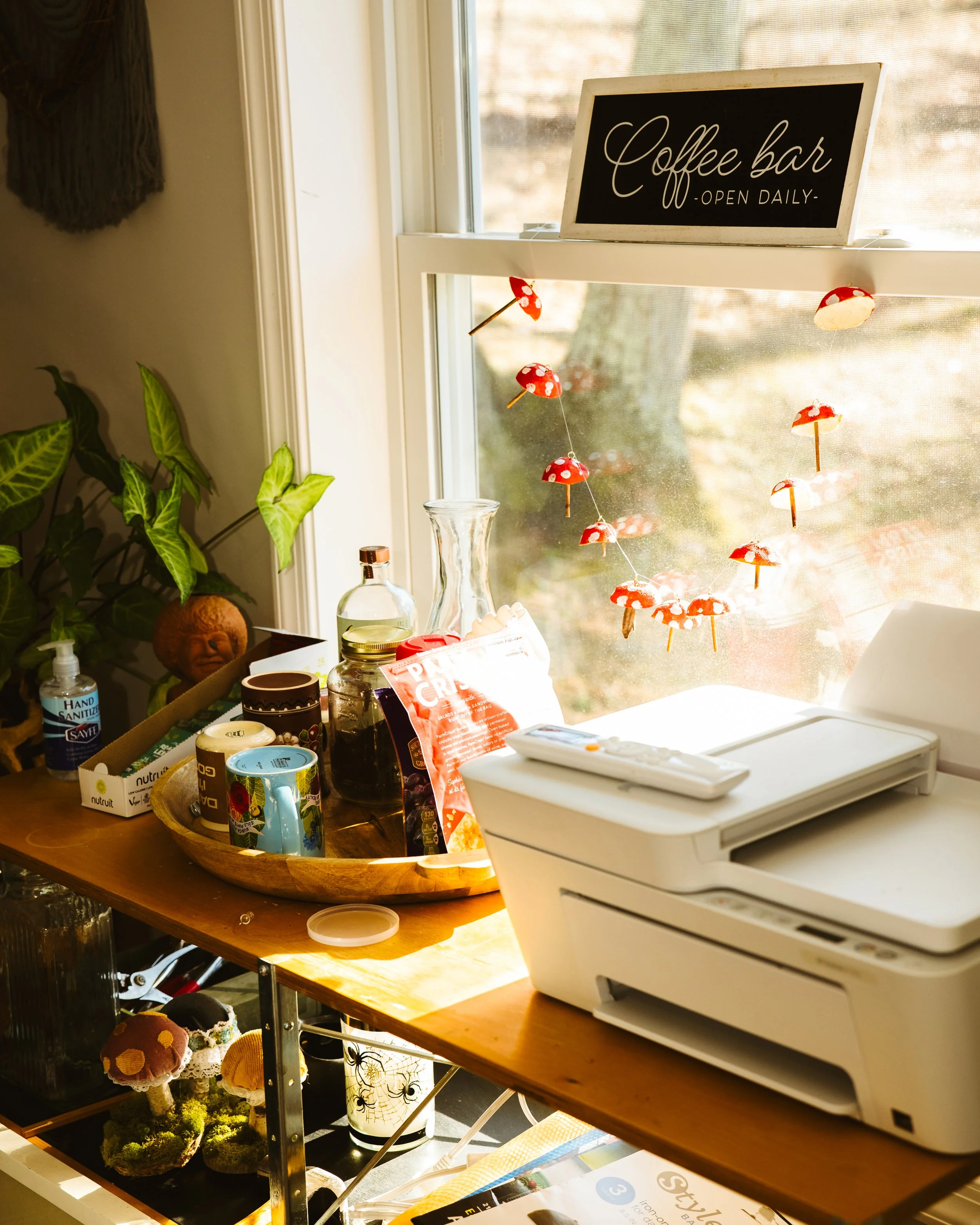 A coffee bar setup next to a window with a sign that reads 'Coffee bar open daily'. Several small mushroom decorations hang from string in the window. Items on the wooden counter include a glass pitcher, a tray with various mugs, a bag of crisps, a p