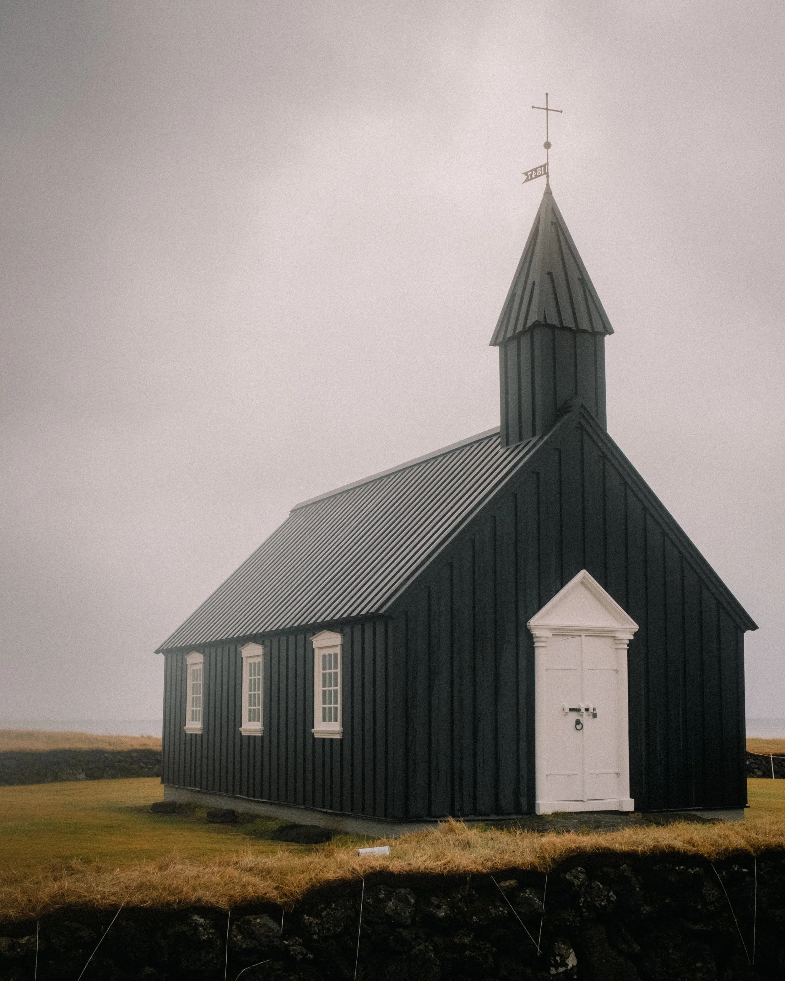 Iceland. Small black wooden chapel with white windows and door, set on a grassy landscape under a cloudy sky. Virginia Travel Photographer. 