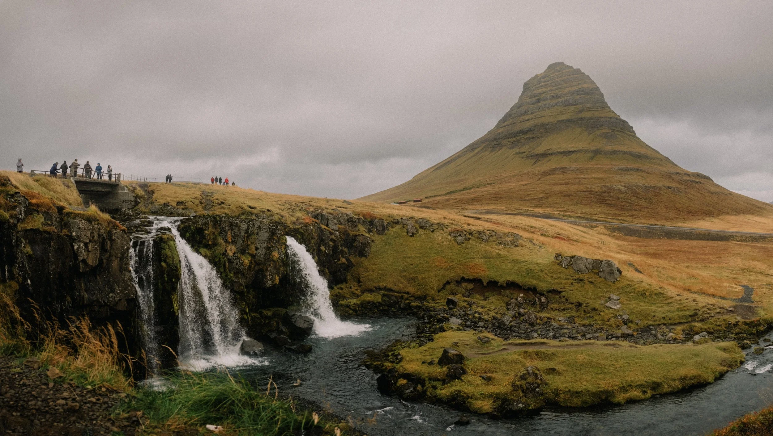 Scenic view of Kirkjufell mountain in Iceland with a small waterfall in the foreground and cloudy sky overhead. Tourists are walking on a pathway near the waterfall. Virginia Travel Photographer.