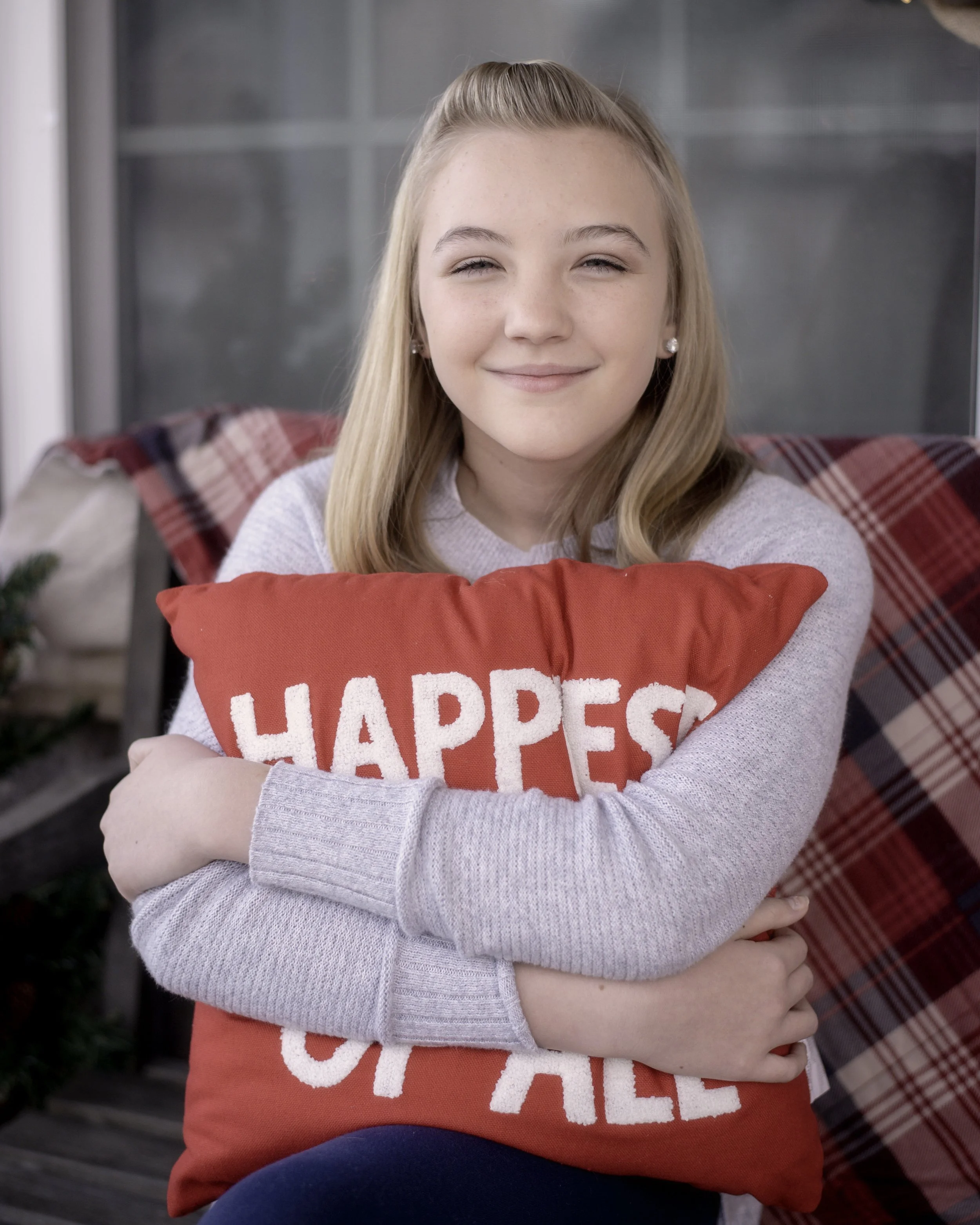 Portrait. A young girl with blonde hair, smiling and hugging a red pillow with the words 'HAPPIEST' and 'OF ALL' written on it, sitting on a porch with plaid cushions in the background. Winchester Portrait Photographer. 