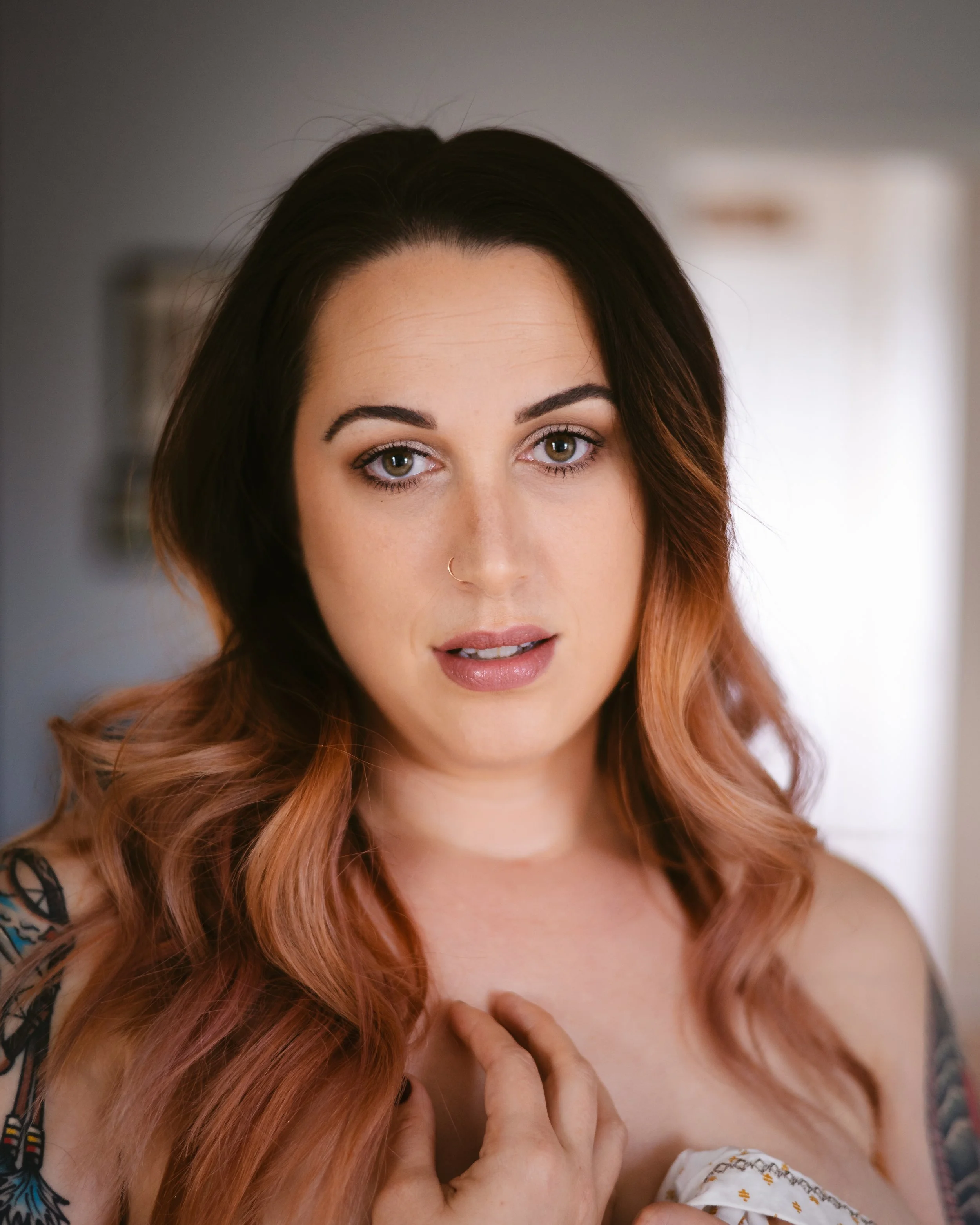 Portrait of a woman. Winchester Portrait Photographer. A woman with long, wavy, pinkish hair and a nose ring, looking into the camera with a soft expression, holding her chest gently with her hand.