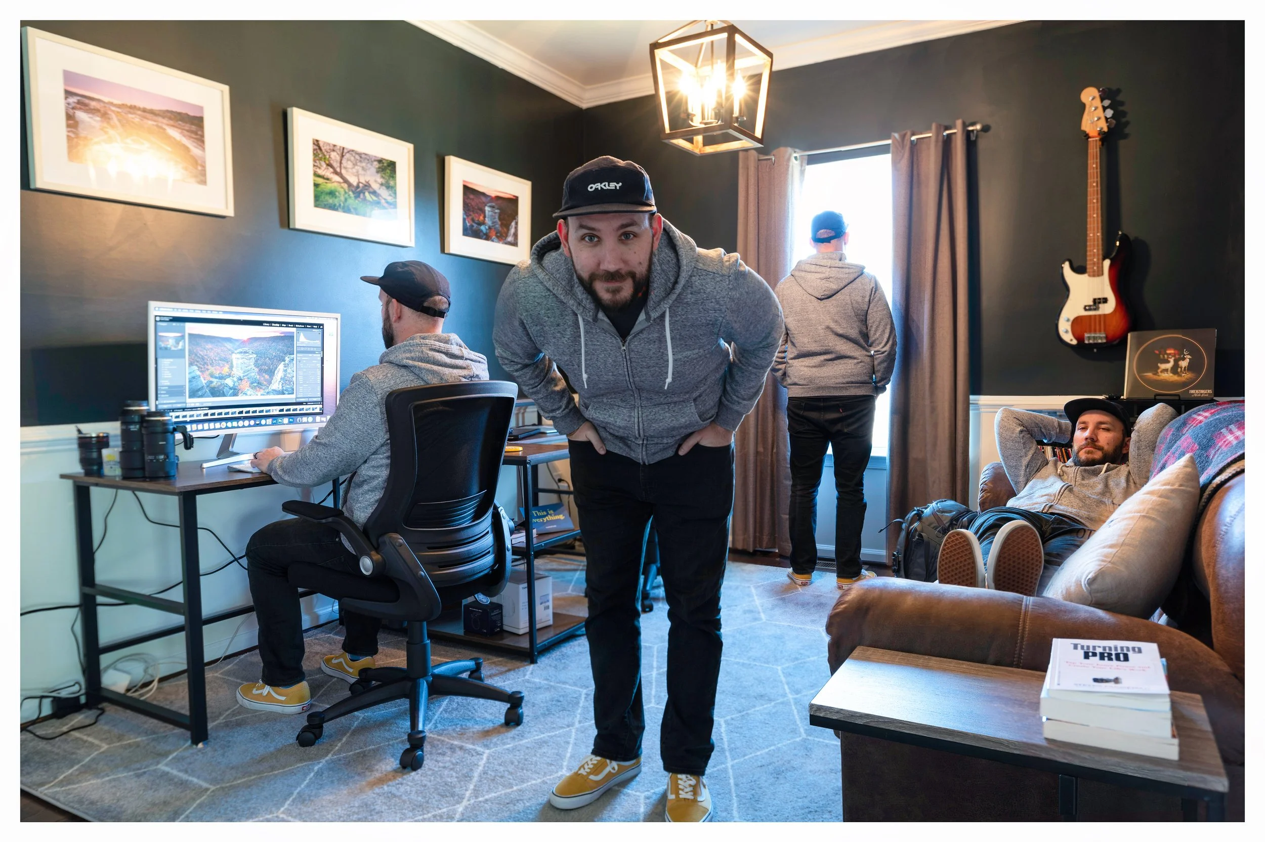 A man in a gray hoodie and black cap bowing towards the camera in a cozy room with four other men. One is working on a computer, another is looking out the window, and two are relaxing on a sofa, one with his feet up and the other leaning back. The room has framed photos, a guitar on the wall, a bookshelf, and a book titled "Turning Pro" on a side table.