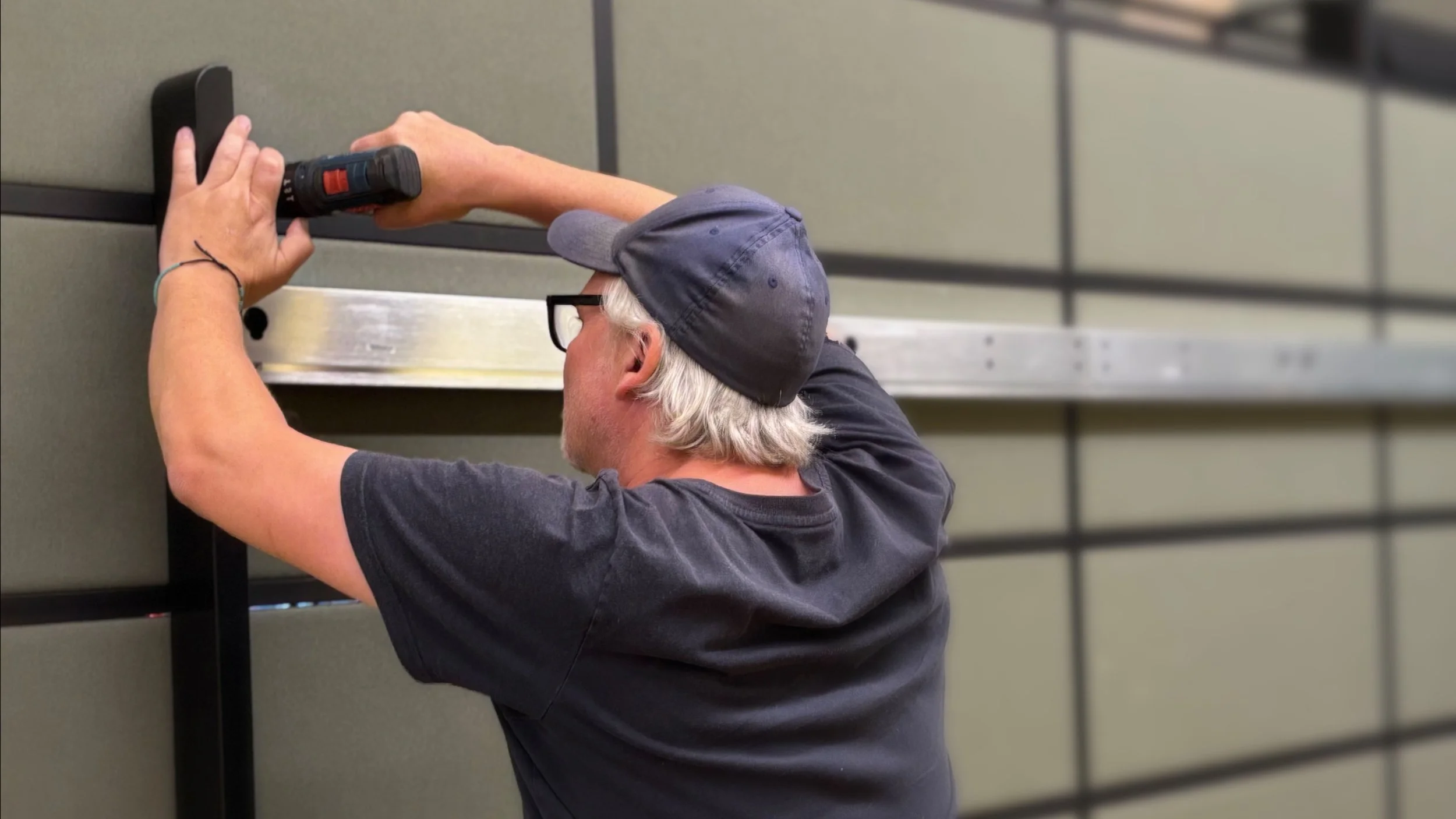 Ein älterer Mann mit grauem Haar, Brille und schwarzem T-Shirt schraubt an einer schwarzen Metallkonstruktion an einer grauen Wand, während er eine Bohrmaschine benutzt.