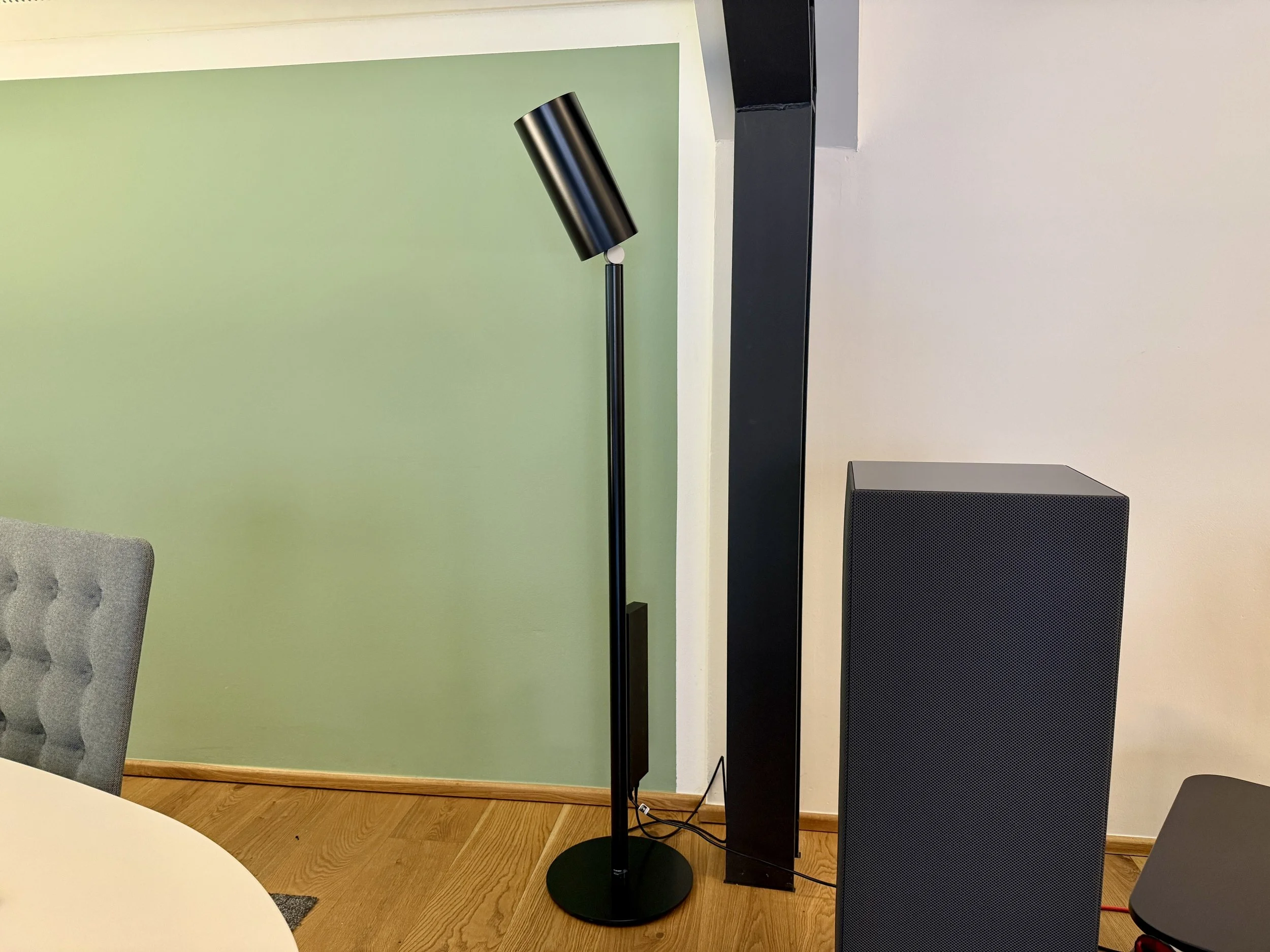 Ein modernes Lautsprechersystem mit einem schwarzen Standlautsprecher und einem schwarzen Bodenrekorder, neben einem schwarzen Subwoofer, in einem Raum mit hellem Holzfußboden und einer grünen Wand.