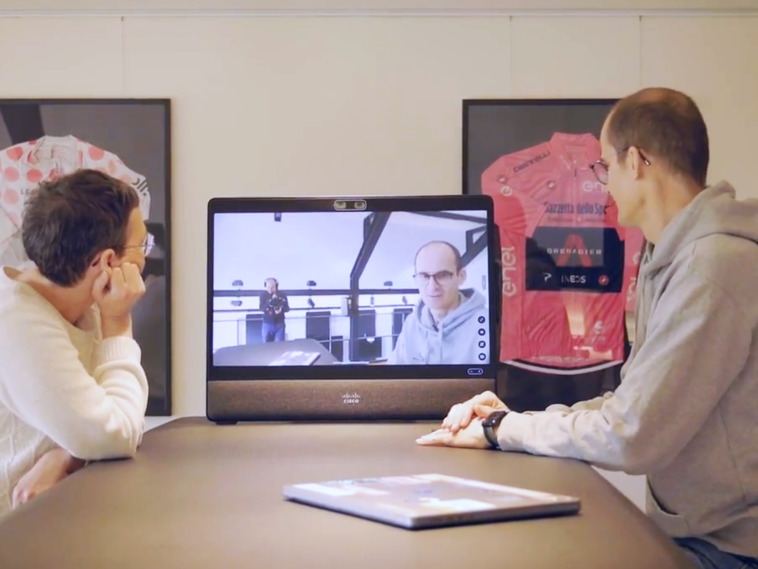 Zwei junge Männer sitzen an einem Tisch und schauen auf einen Bildschirm, der eine Person zeigt, die auf einem Balkon steht.