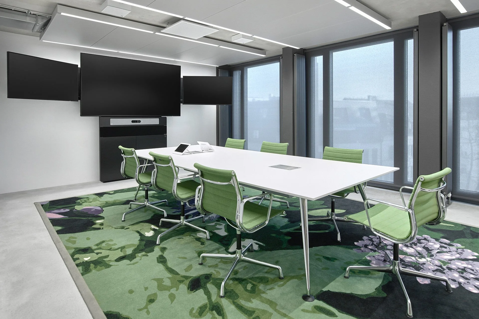 Ein Moderner Konferenzraum mit weißen Tischen, grünen Stühlen, großen Fenstern mit Vorhängen, einem großen Bildschirm an der Wand und einer grünen Teppich mit Blumenmuster.