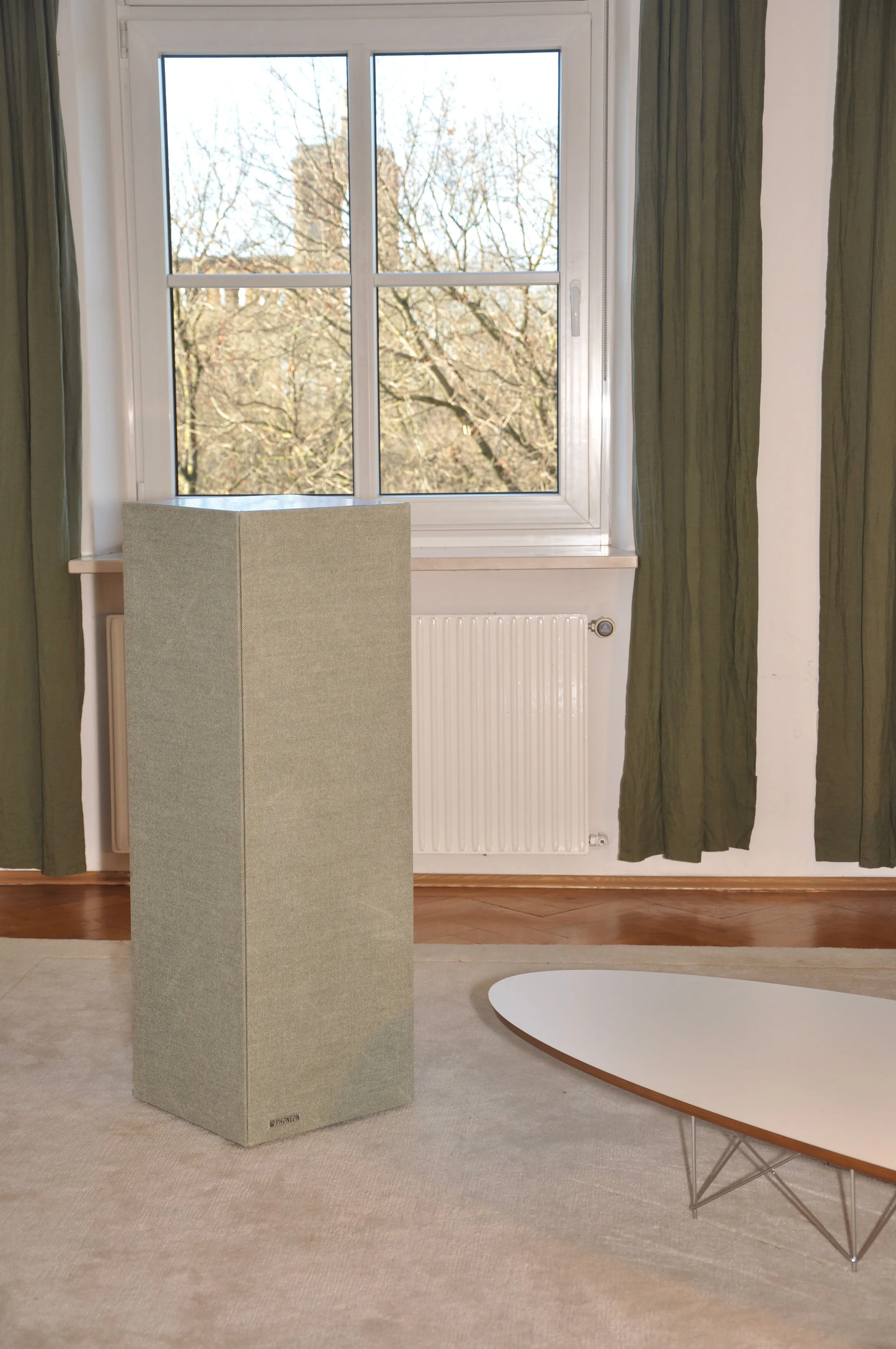 Ein Raum mit einem großen Fenster, grünen Vorhängen, einem weißen Radiator, einer großen neutralfarbenen Lautsprecherbox und einem modernen, ovalen Couchtisch auf einem beige Teppichboden.