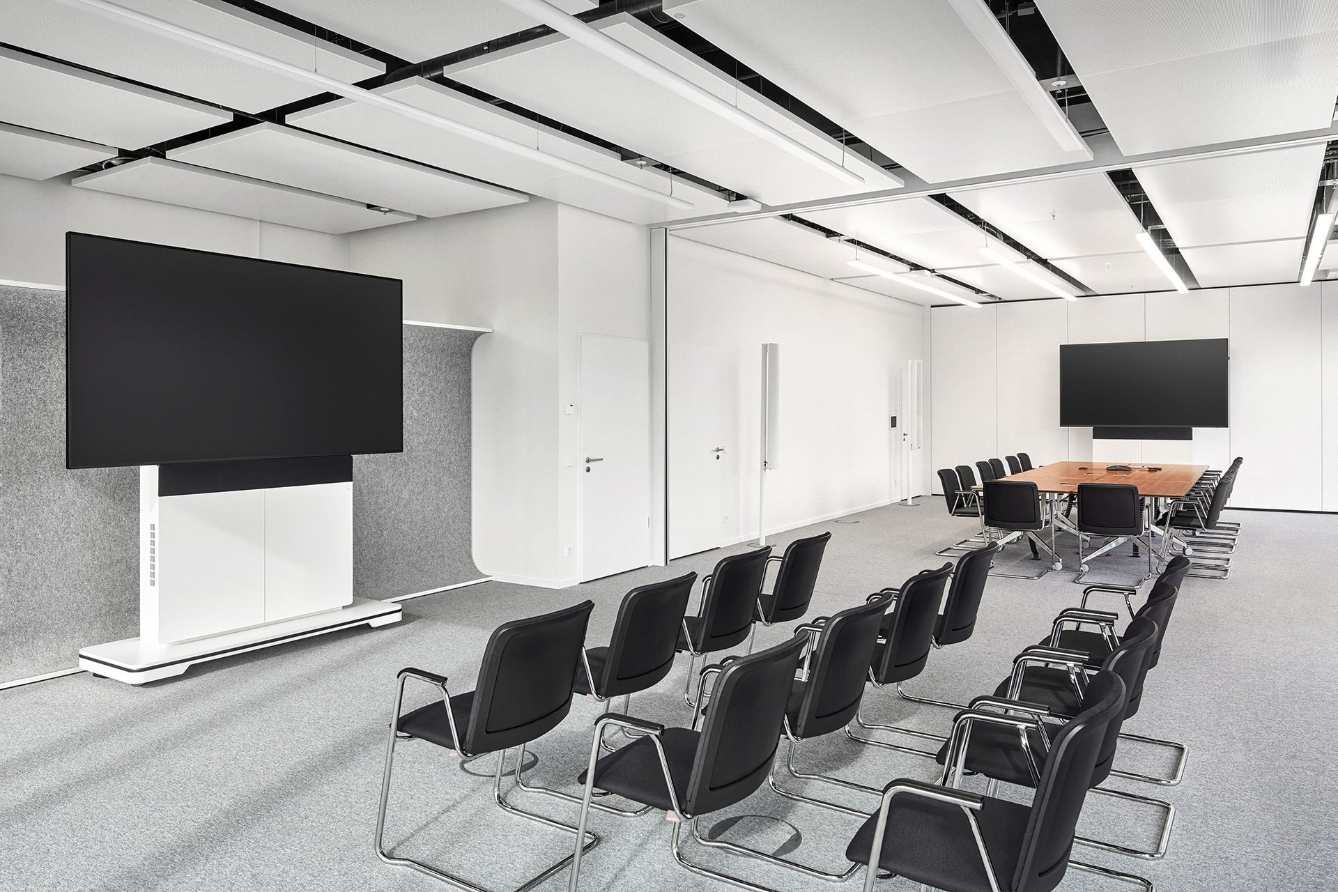 Ein moderner, leerer Konferenzraum mit Stühlen, zwei große Bildschirme, einem langen Holztisch und weißen Wänden.
