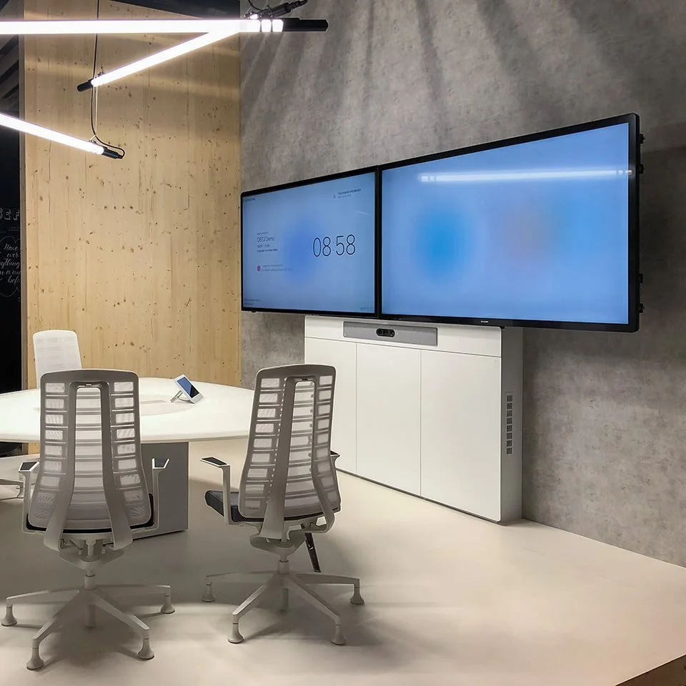 Moderner Konferenzraum mit weißen Tischen, ergonomischen Stühlen und zwei großen digitalen Displays an der Wand.