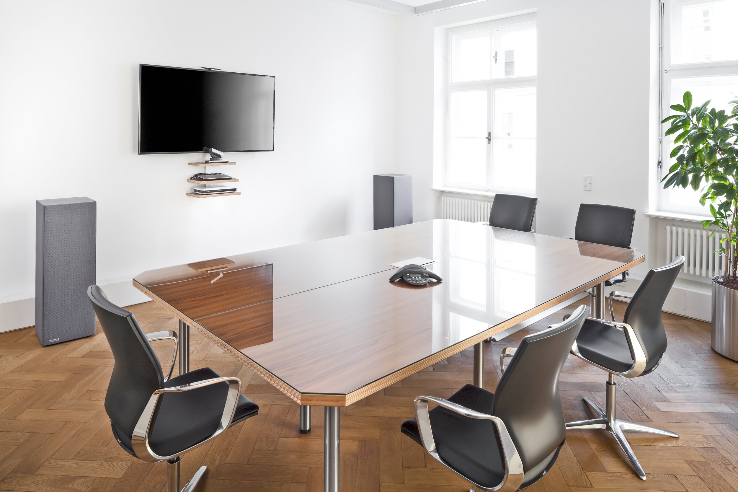 Ein modernes Konferenzraum mit einem großen Holztisch, sechs schwarzen Stuhl, einem großen Flachbildschirm an der Wand, einer an der Wand montierten Konferenztelefon Konferenztelefon, zwei große Fenster, eine Topfpflanze in einem Metalltopf.