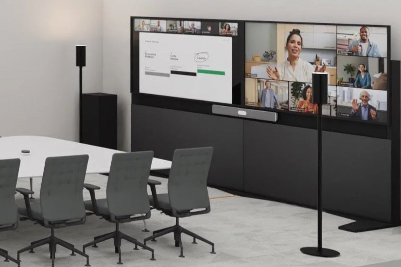 Ein Konferenzraum mit einem großen Bildschirm, auf dem eine Video-Konferenz mit mehreren Teilnehmern läuft, und einem weißen Tisch mit Stühlen.