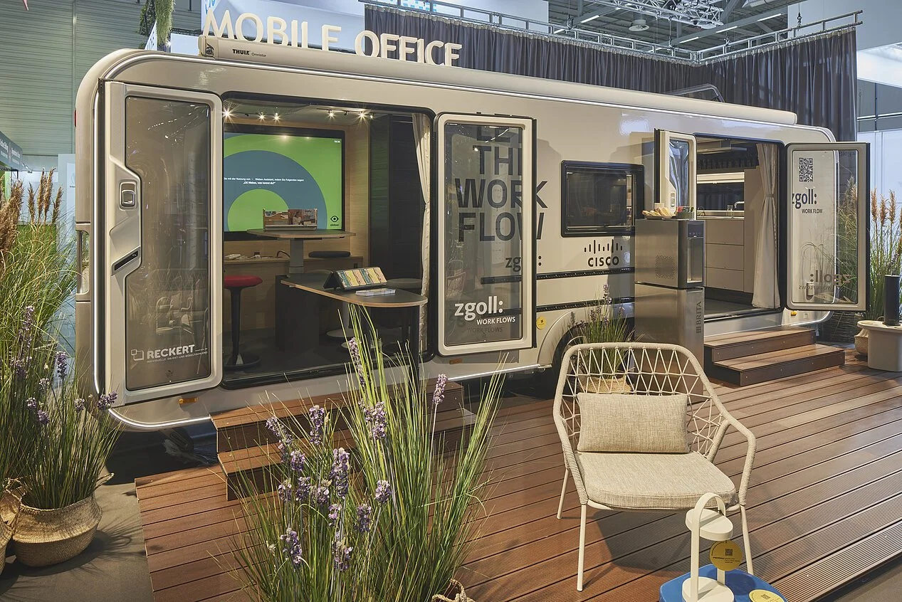 Ein umgebauter Wohnwagen als mobiles Büro mit offener Tür, Holzdecke, Pflanzen im Vordergrund und einem bequemen Gartenstuhl auf Holzdeck. Innen sind mehrere Monitore und Büromöbel sichtbar.