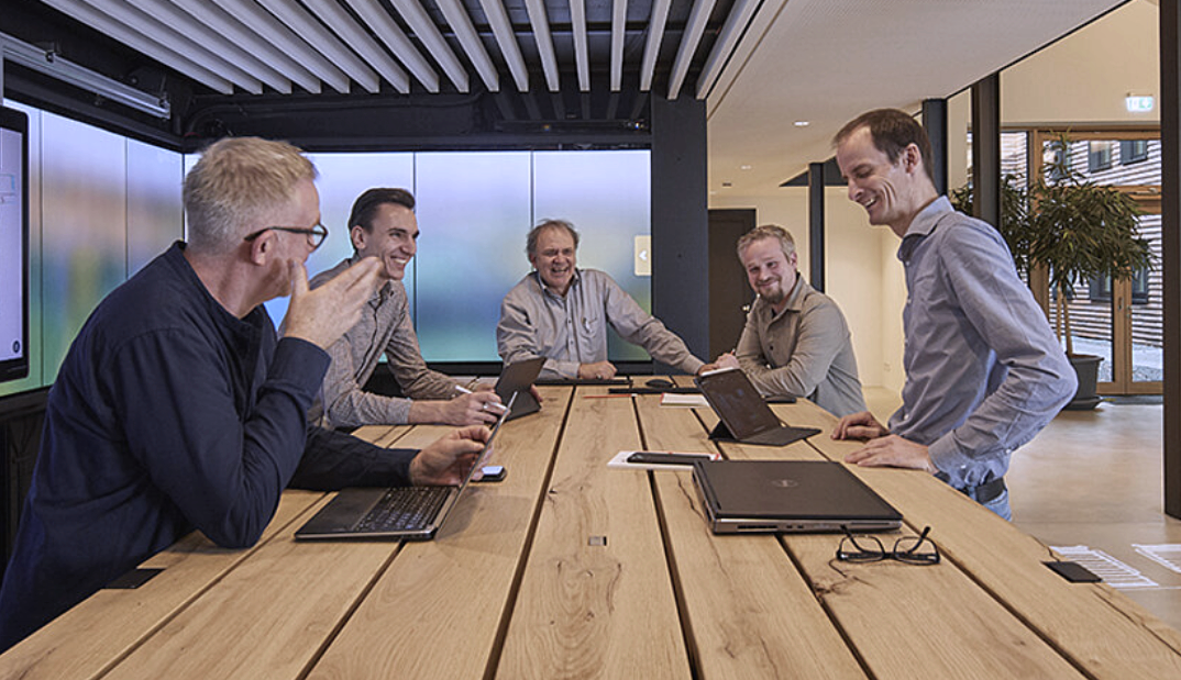 Fünf Männer sitzen und stehen an einem Konferenztisch in einem modernen Meetingraum, lachen und unterhalten sich, mit Laptops und Tablets vor sich.