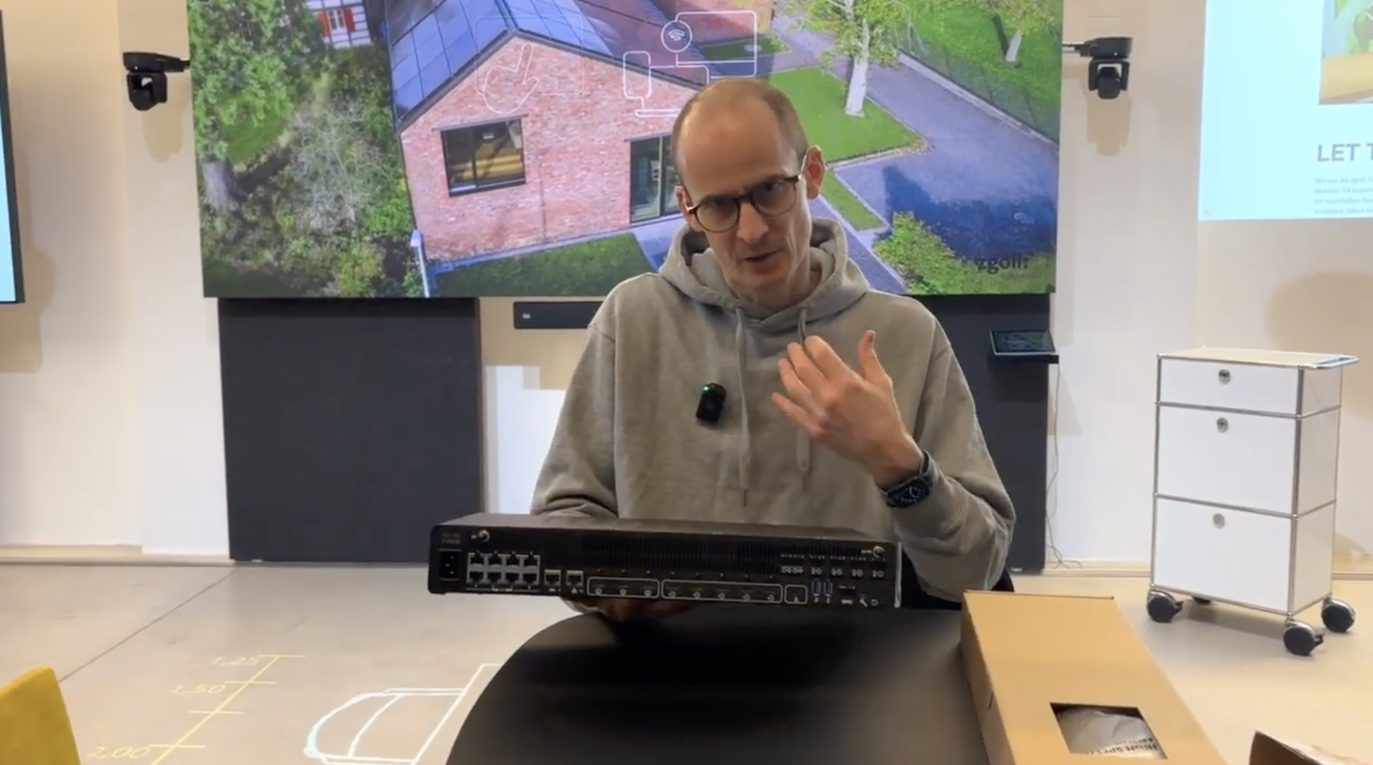 Mann mit Brille und grauer Kapuzenjacke sitzt an einem Tisch mit einer Netzwerk-Switch, in einem Raum mit großen Bildschirmen im Hintergrund.