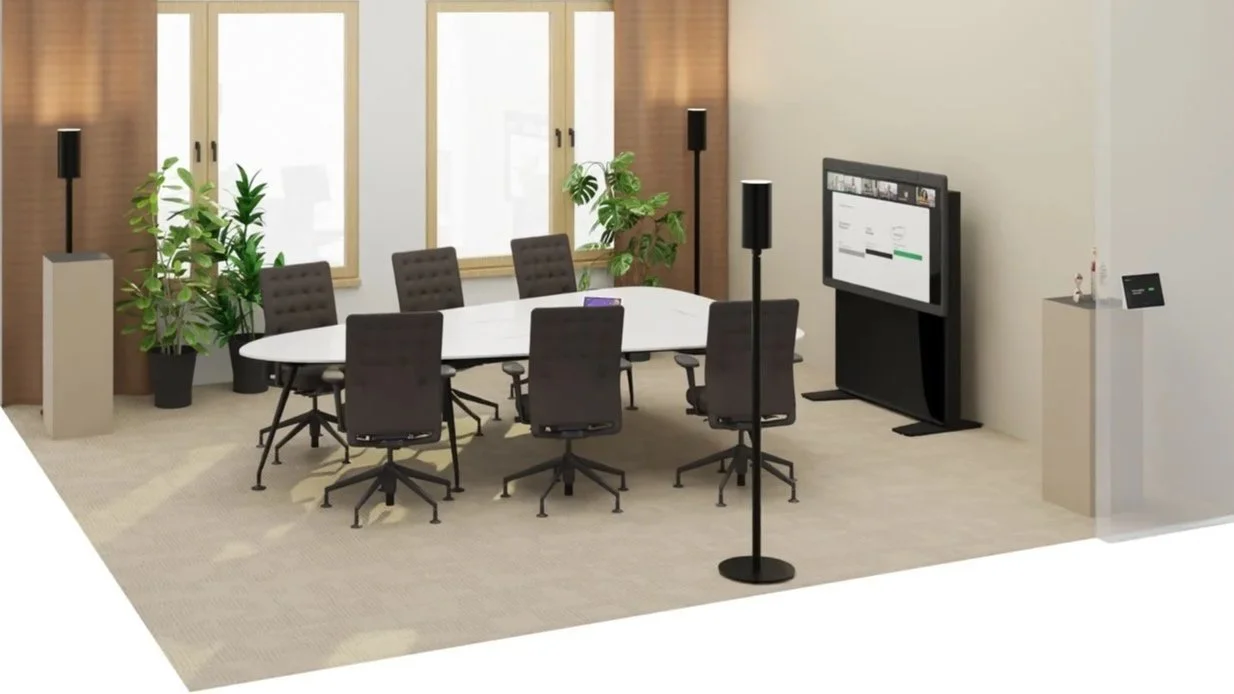 Konferenzraum mit einem ovalen Tisch, sechs Stühlen, Pflanzen, einem großen Bildschirm, Lautsprechern und einem Gerät auf einem Podest.