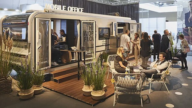 Ein mobiles Büro in einem Bus, umgeben von Menschen, die sich unterhalten und entspannen, mit Pflanzen im Vordergrund.