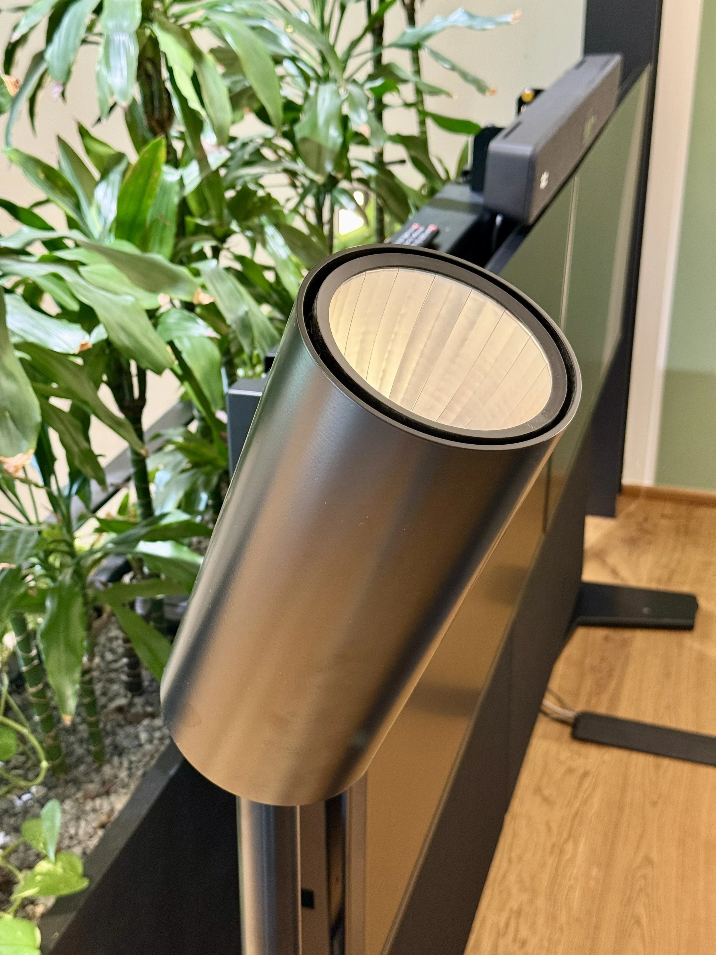 Ein moderner, silberner Luftreiniger steht auf einem schwarzen Möbelstück neben einer Pflanze mit großen grünen Blättern. Im Hintergrund befinden sich ein Telefon und eine Wand.