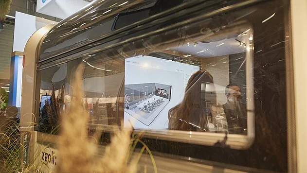 Blick durch eine große Glasscheibe auf eine ausgestellte technische Maschine mit einem digitalen Bildschirm im Hintergrund, im Vordergrund Pflanzen und Unschärfe.