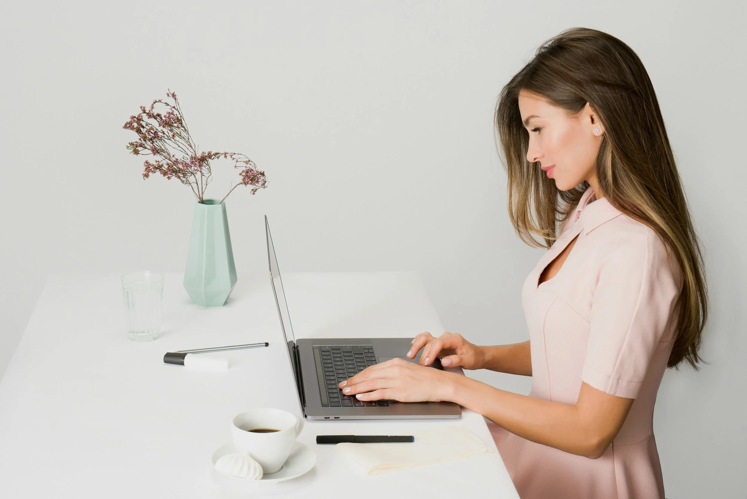 Woman working on laptop at a white table with a vase of flowers, a glass of water, a cup of coffee, and writing utensils.