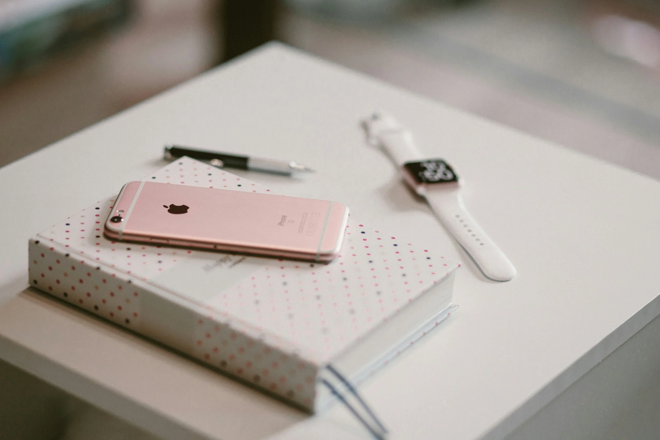 A white table with a pink iPhone, a black and white pen, a wrapped gift box with red and black polka dots, and a smartwatch with a white band and black face.