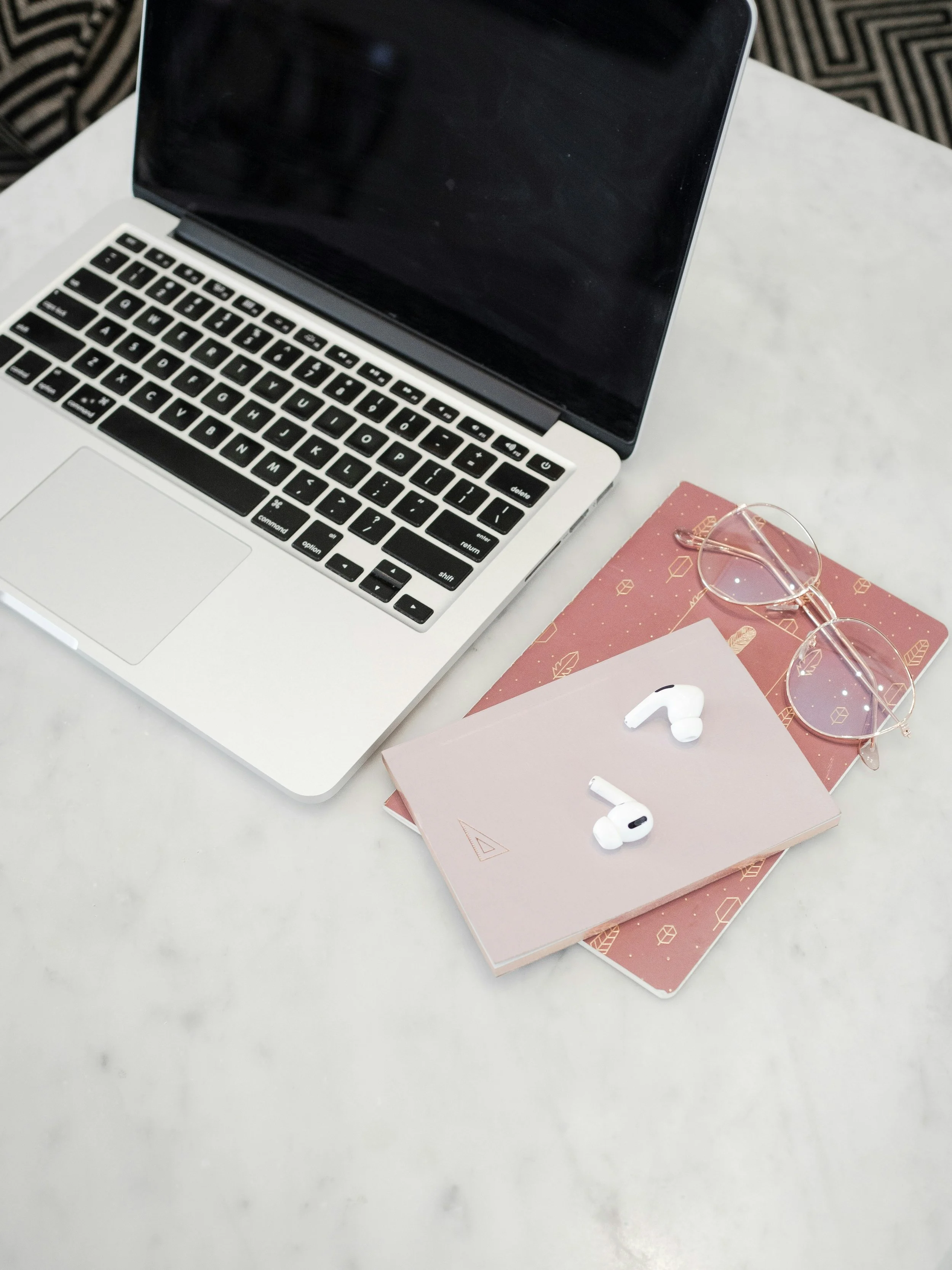 A silver laptop, a pair of pink glasses, two notebooks (pink with gold geometric designs and light pink), and white wireless earbuds on a white marble surface.