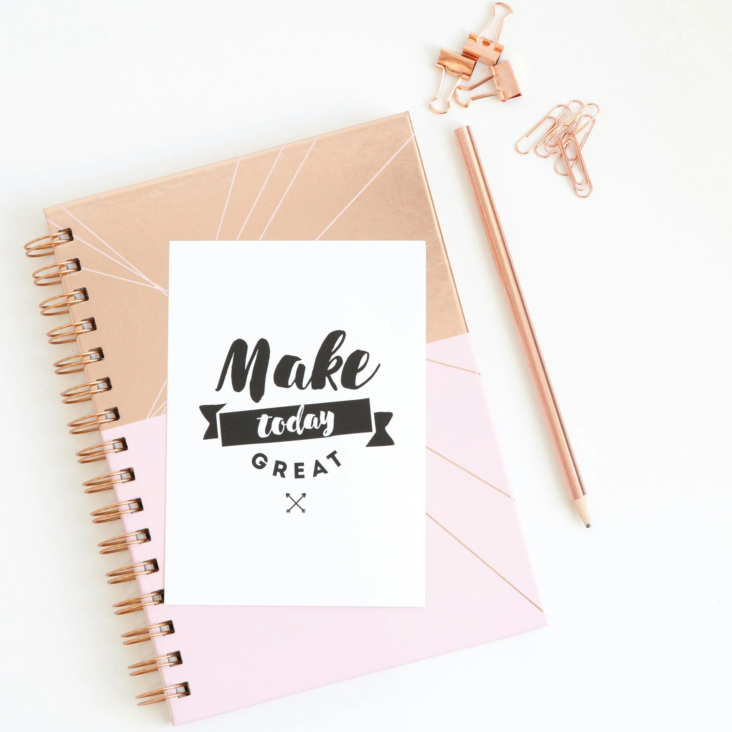 A pink and rose gold spiral notebook with a motivational quote card that reads 'Make today great,' accompanied by rose gold paper clips, binder clips, and a ballpoint pen on a white surface.