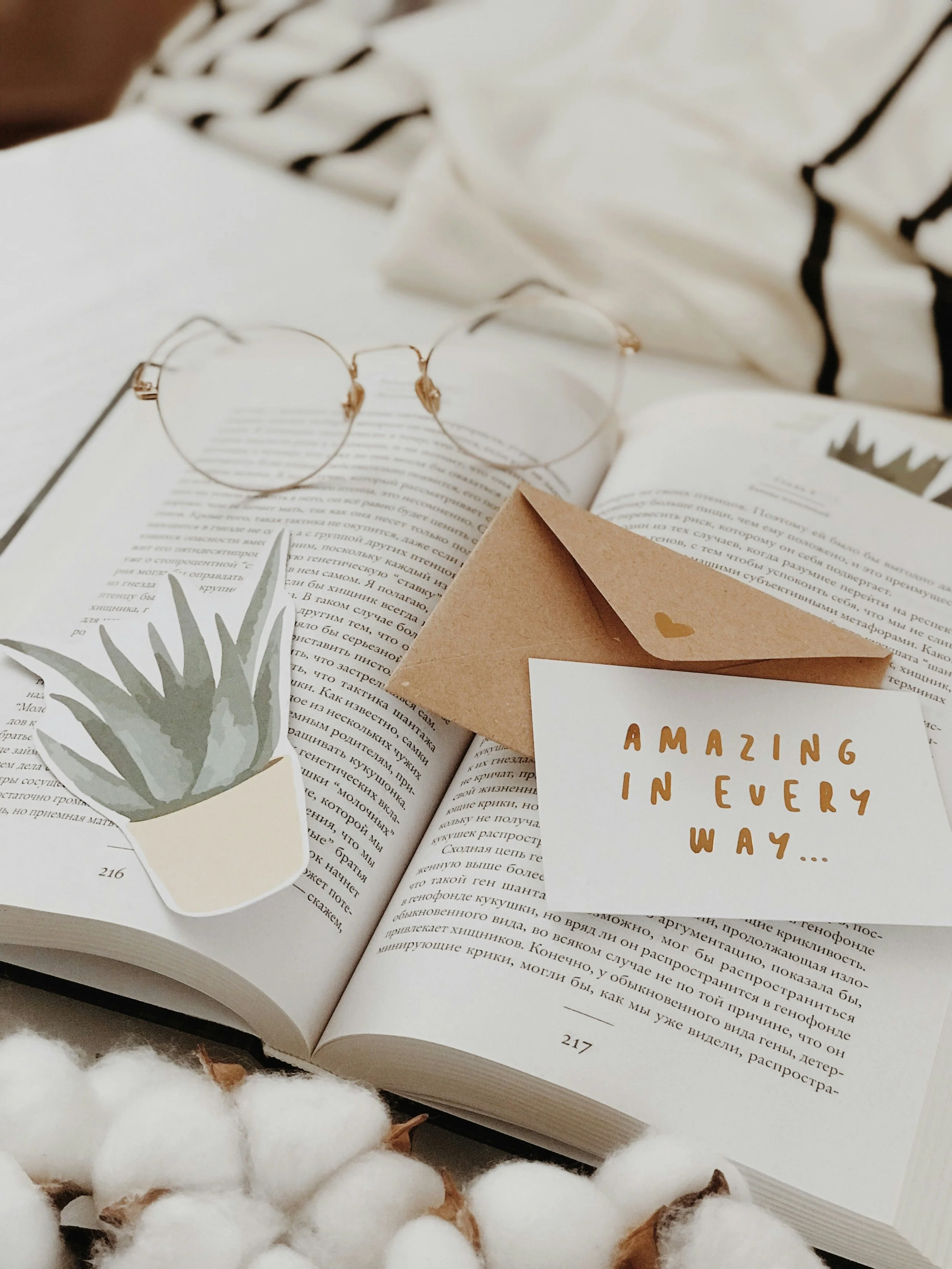 Open book with glasses, a small potted plant illustration, a brown paper envelope, and a card with the text 'AMAZING IN EVERY WAY' on it. Cotton flowers are in the foreground.