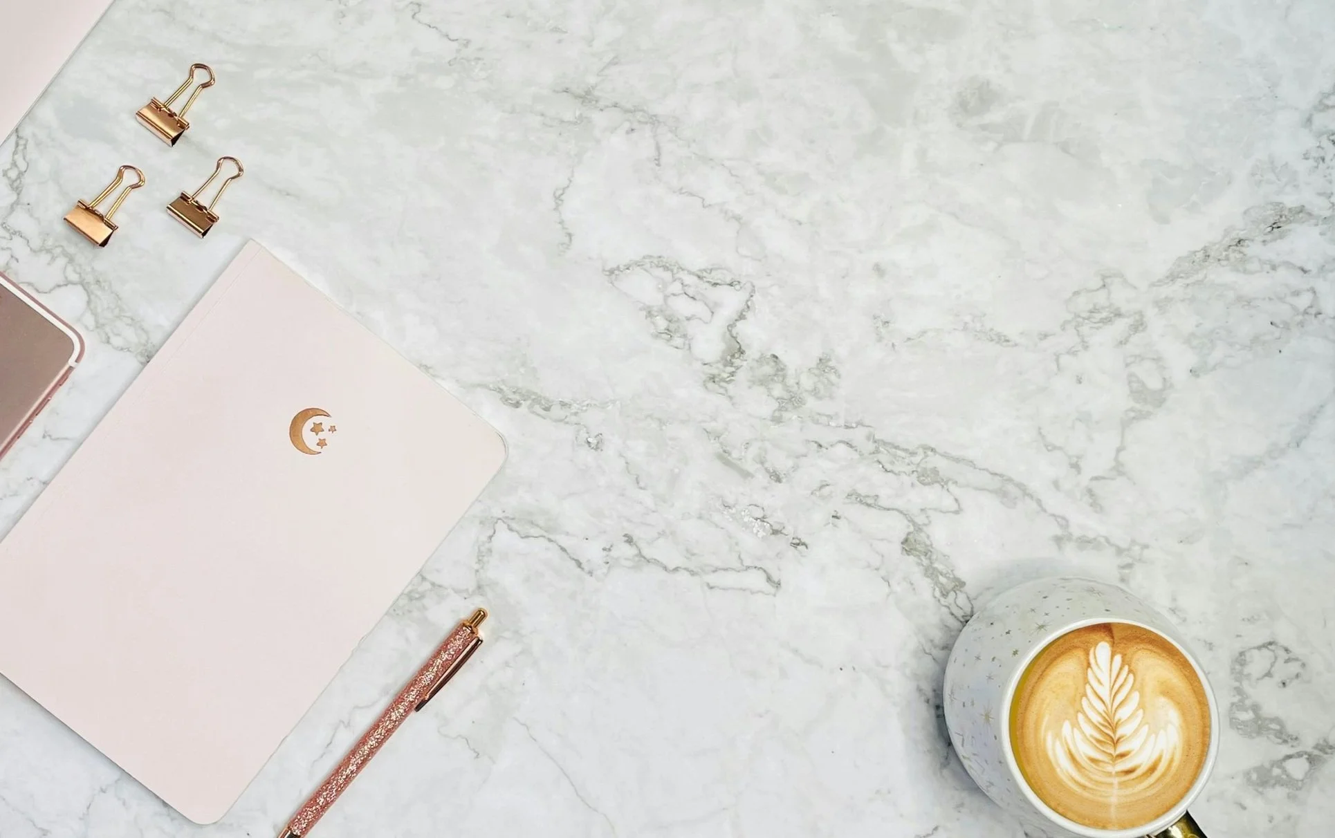 A marble surface with three gold binder clips, a pink pen, a white notebook with a crescent moon and stars design, a rose gold device, and a cup of latte with latte art.