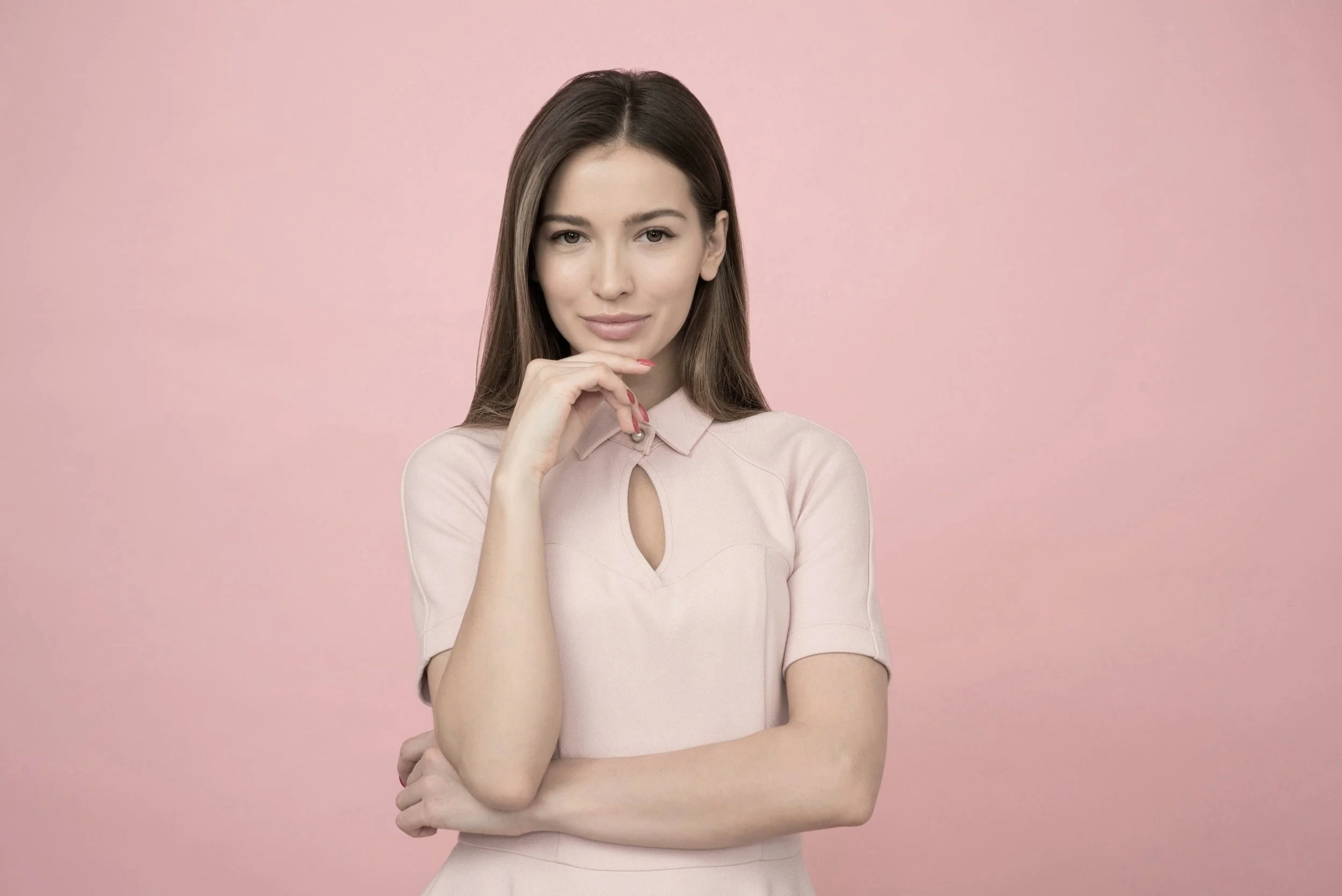 A young woman with long brown hair, wearing a light pink dress with a keyhole neckline, standing against a pink background, smiling subtly at the camera.