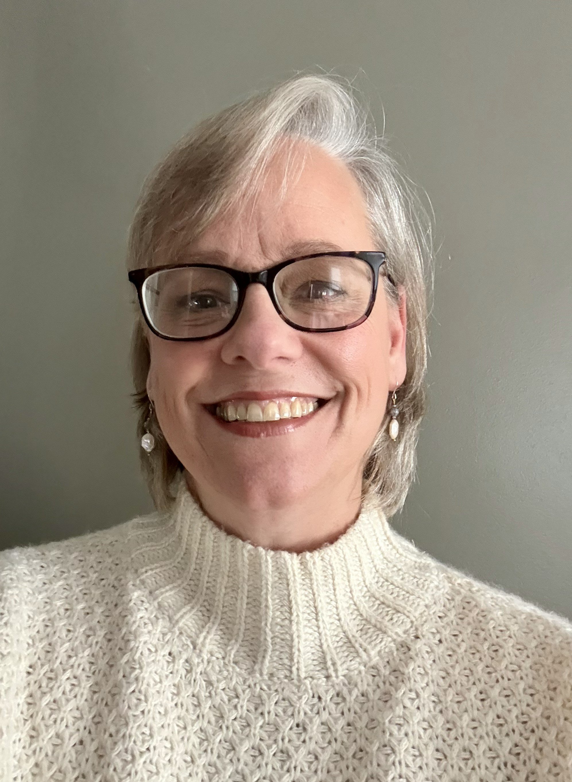 A woman with short gray hair, glasses, and earrings smiling for the camera, wearing a cream-colored knit turtleneck sweater against a plain wall background.