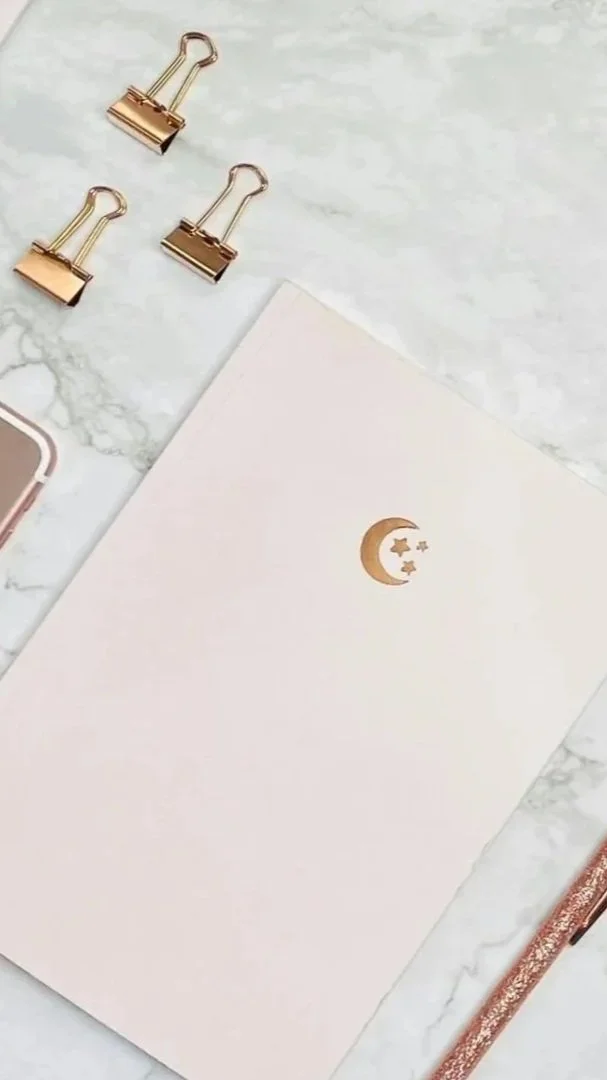 A white notepad with a gold crescent moon and stars design, three gold binder clips, a pen, and part of a smartphone on a marble surface.