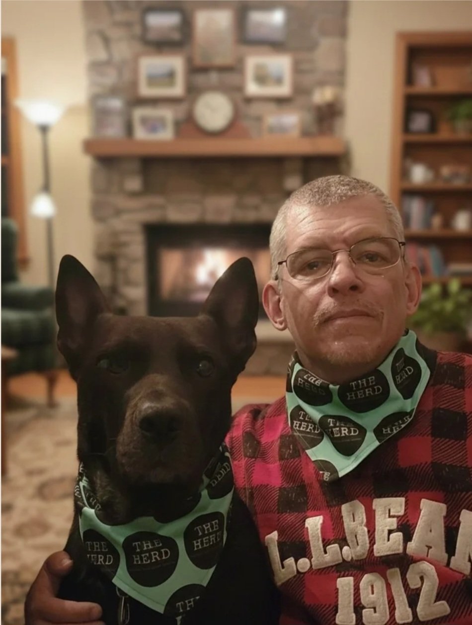 A man with glasses and a dog sitting together in a cozy living room with a fireplace, bookshelf, and pictures on the wall in the background. Both are wearing bandanas with the logo 'The Herd'.