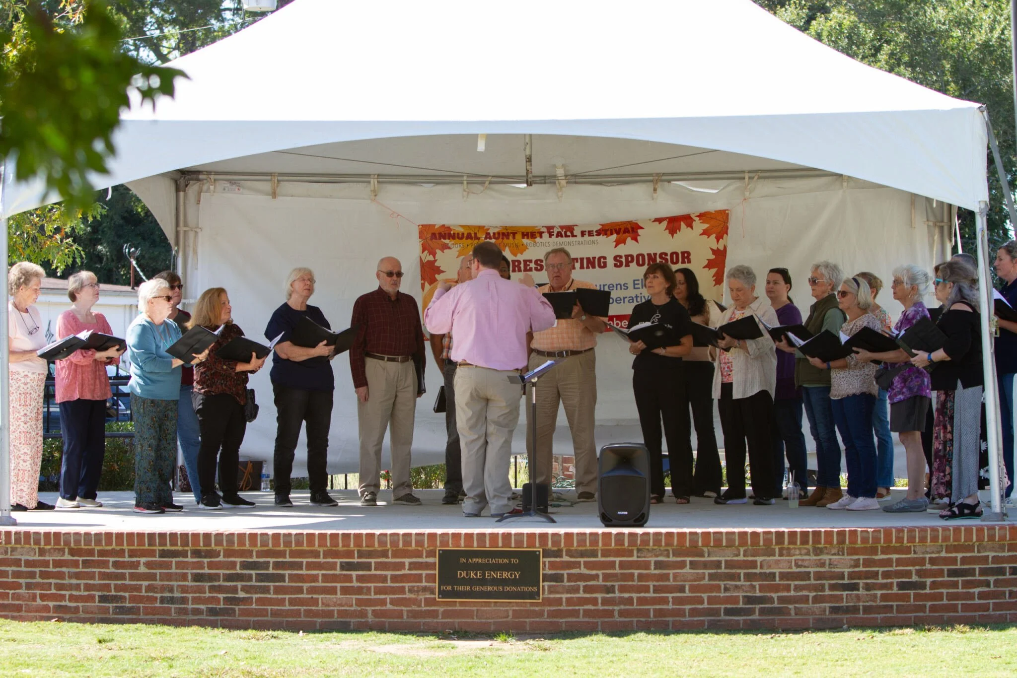 A choir of elderly men and women singing on an outdoor stage at a fall festival, led by a conductor in a pink shirt. The stage is covered with a white canopy and has a banner that reads 'Annual Aunt Het Fall Festival' and 'Celebrating Sponsor'. A plaque at the front of the stage thanks Duke Energy for their donations.