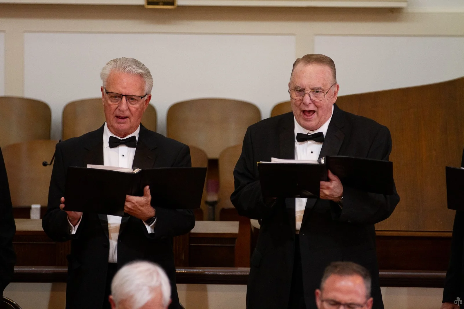 Two elderly men in tuxedos singing in a choir, holding black folders with sheet music, in a church or concert hall.