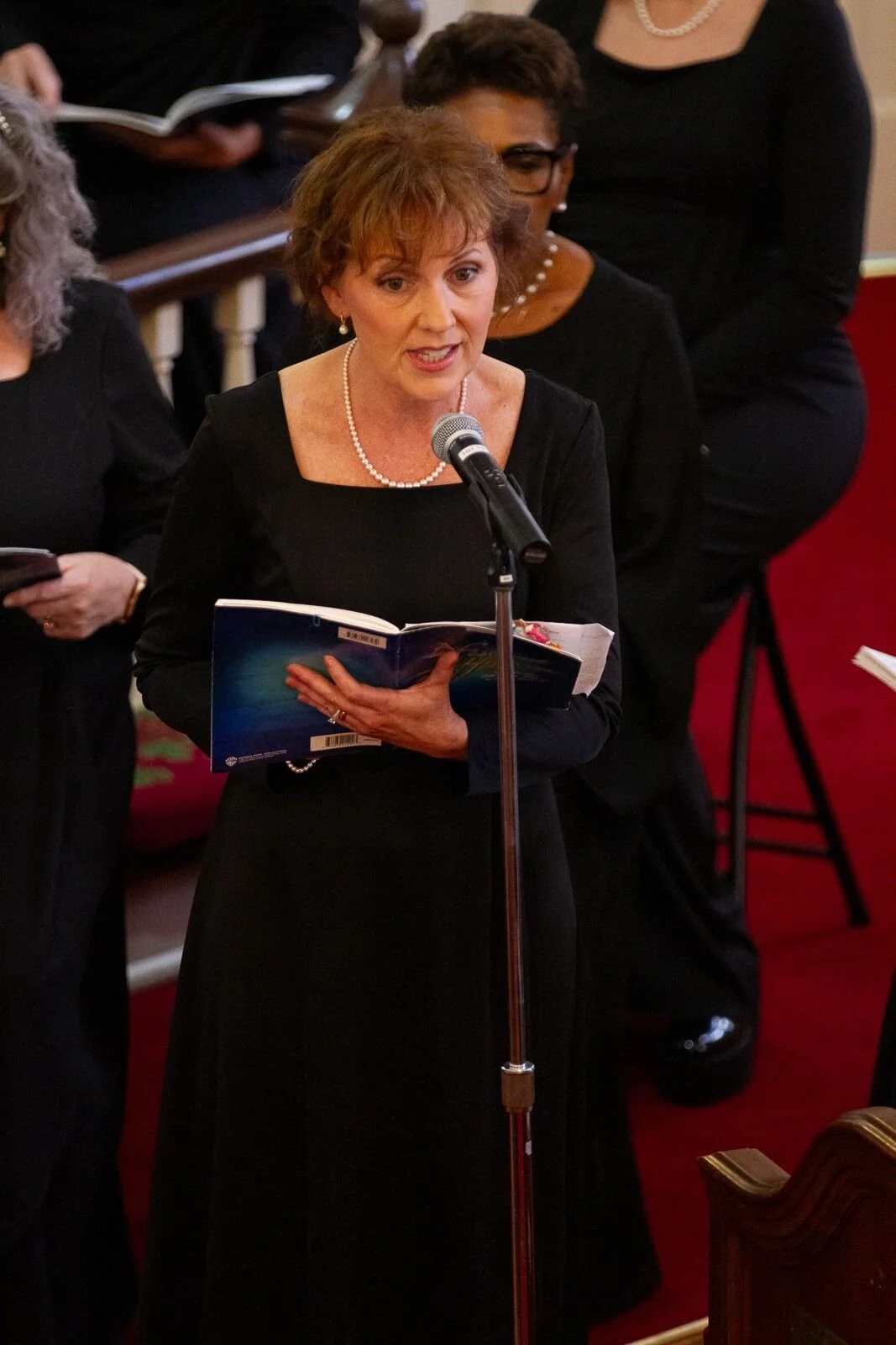 A woman dressed in black, wearing pearl necklace and earrings, speaking into a microphone while holding a book. Several other people in black outfits are visible behind her, some with books or phones in hand.